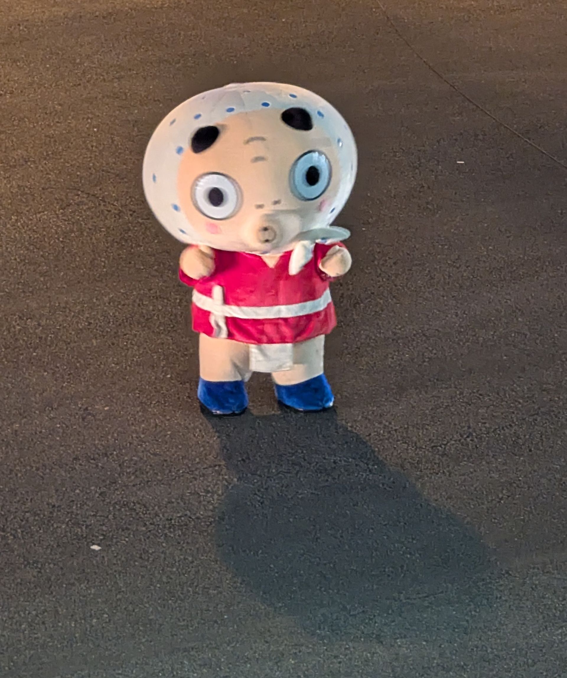 Mascot in red outfit with blue shoes, light skin, on dark asphalt.