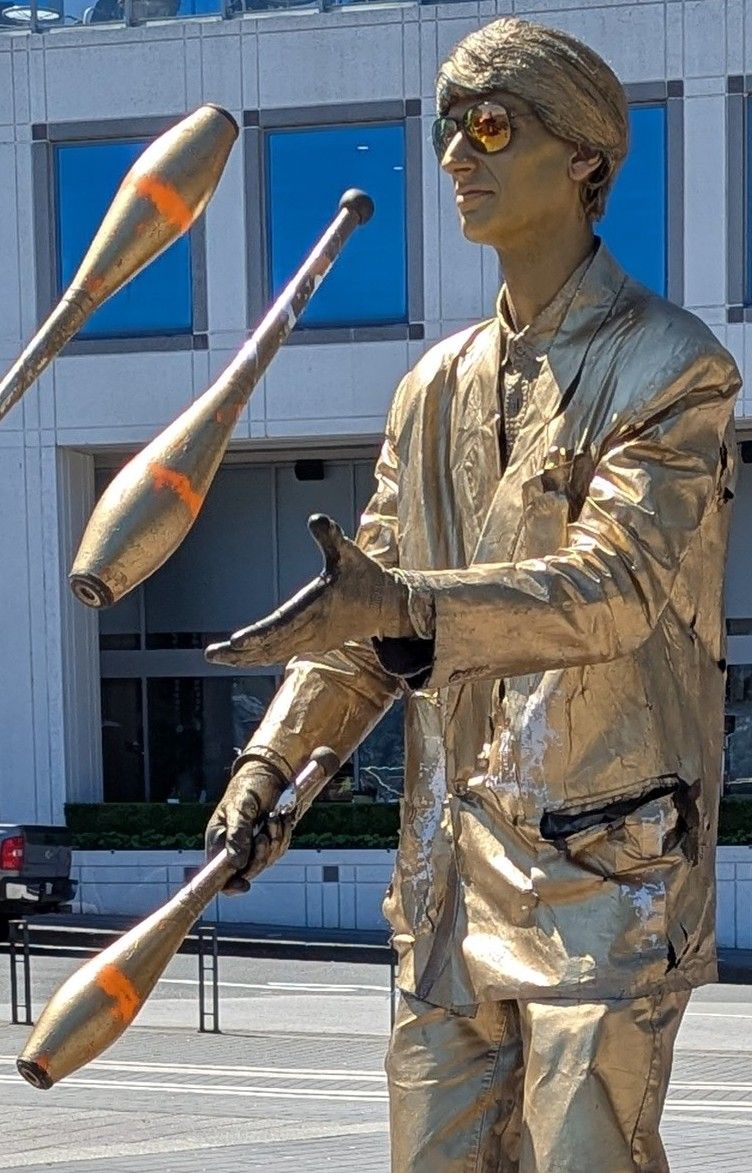A statue of a man painted gold is juggling three juggling pins.