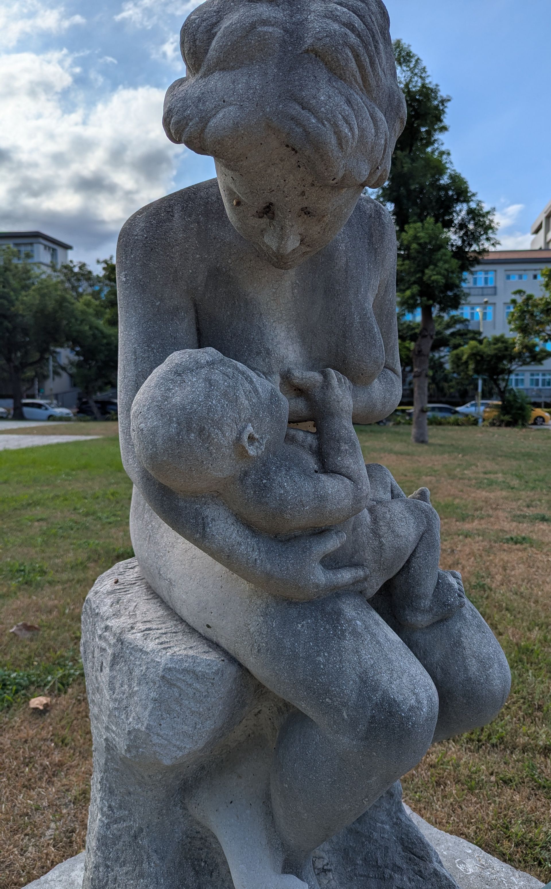 Statue of a mother breastfeeding a baby, seated outdoors. Gray stone sculpture with details of skin and fabric.