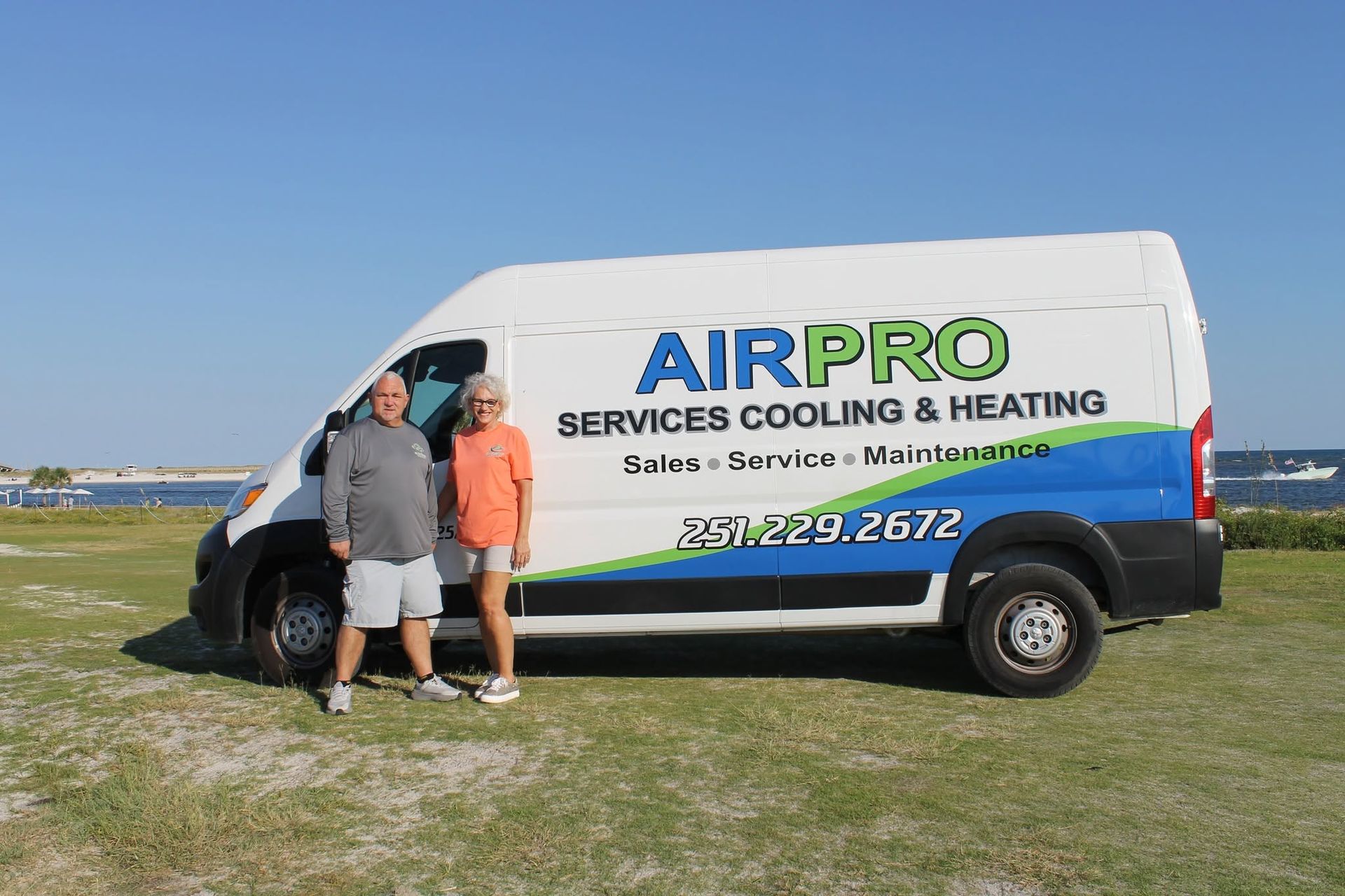 Two people stand by an AIRPRO service van parked on grass under a clear blue sky.