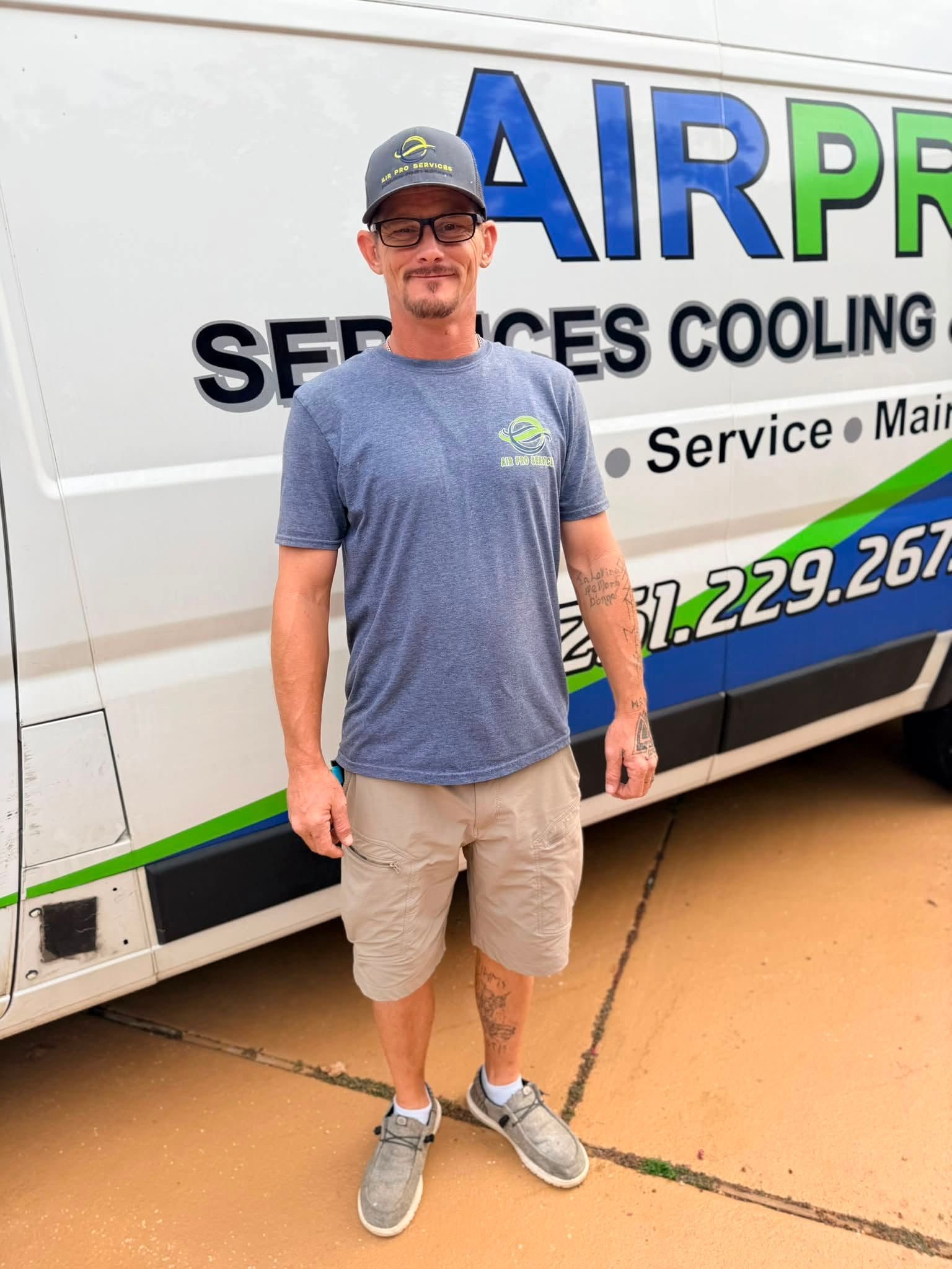 Man standing in front of a white service van with blue and green lettering, smiling and wearing a cap.