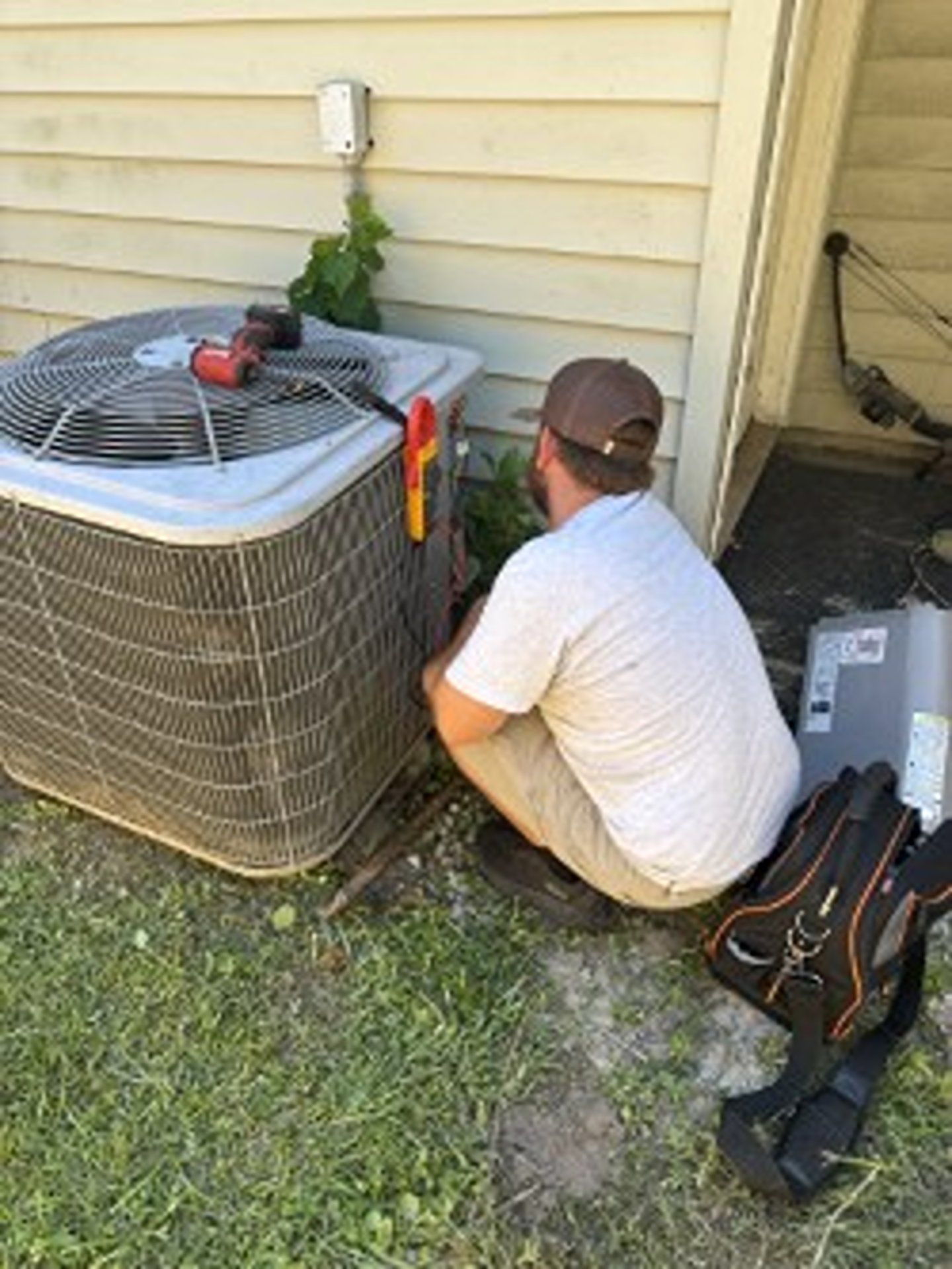 Technician kneeling beside outdoor air conditioner unit, working with tools near house wall