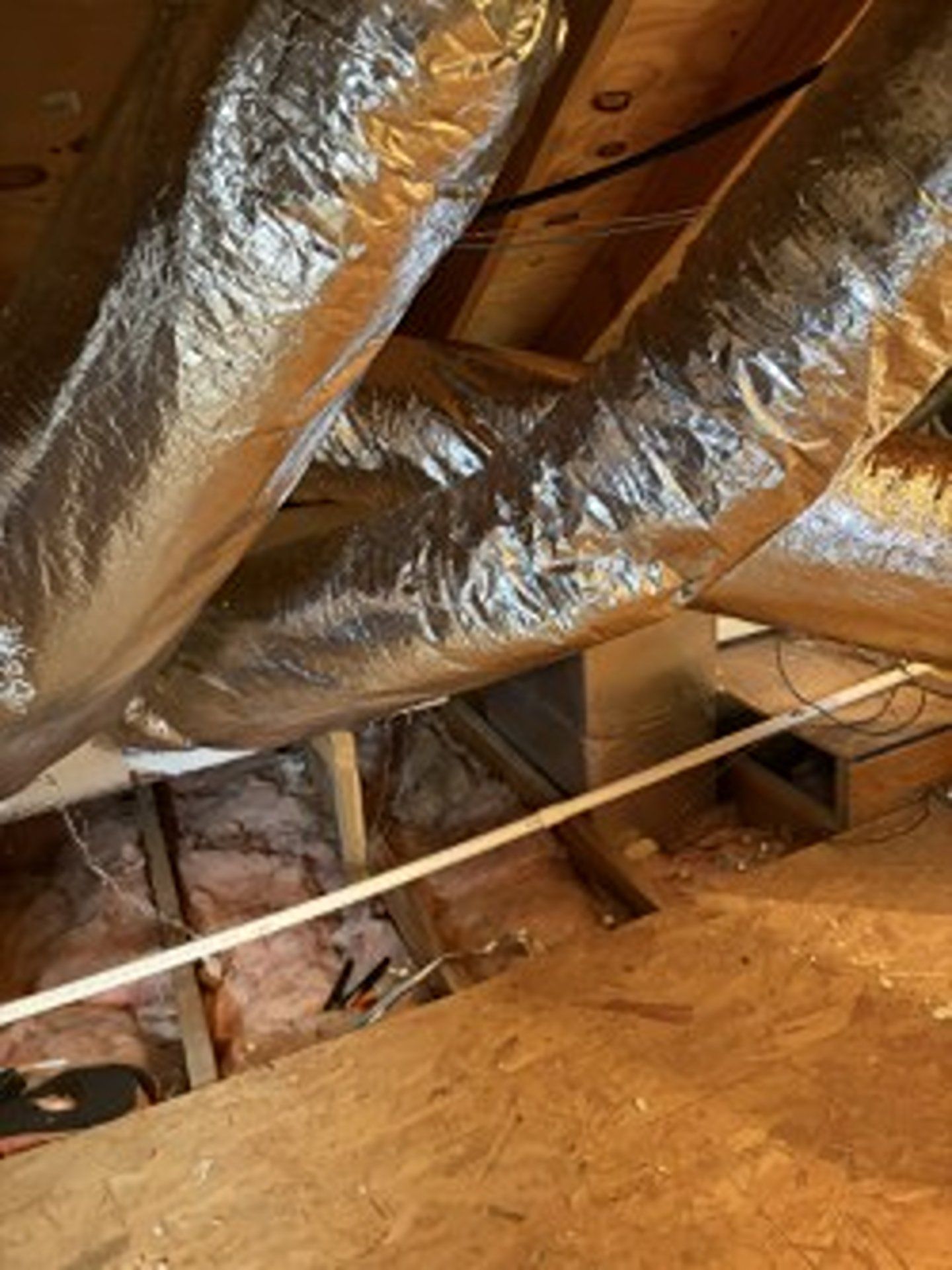 Exposed attic with silver insulated ductwork and wooden beams above a dusty floor