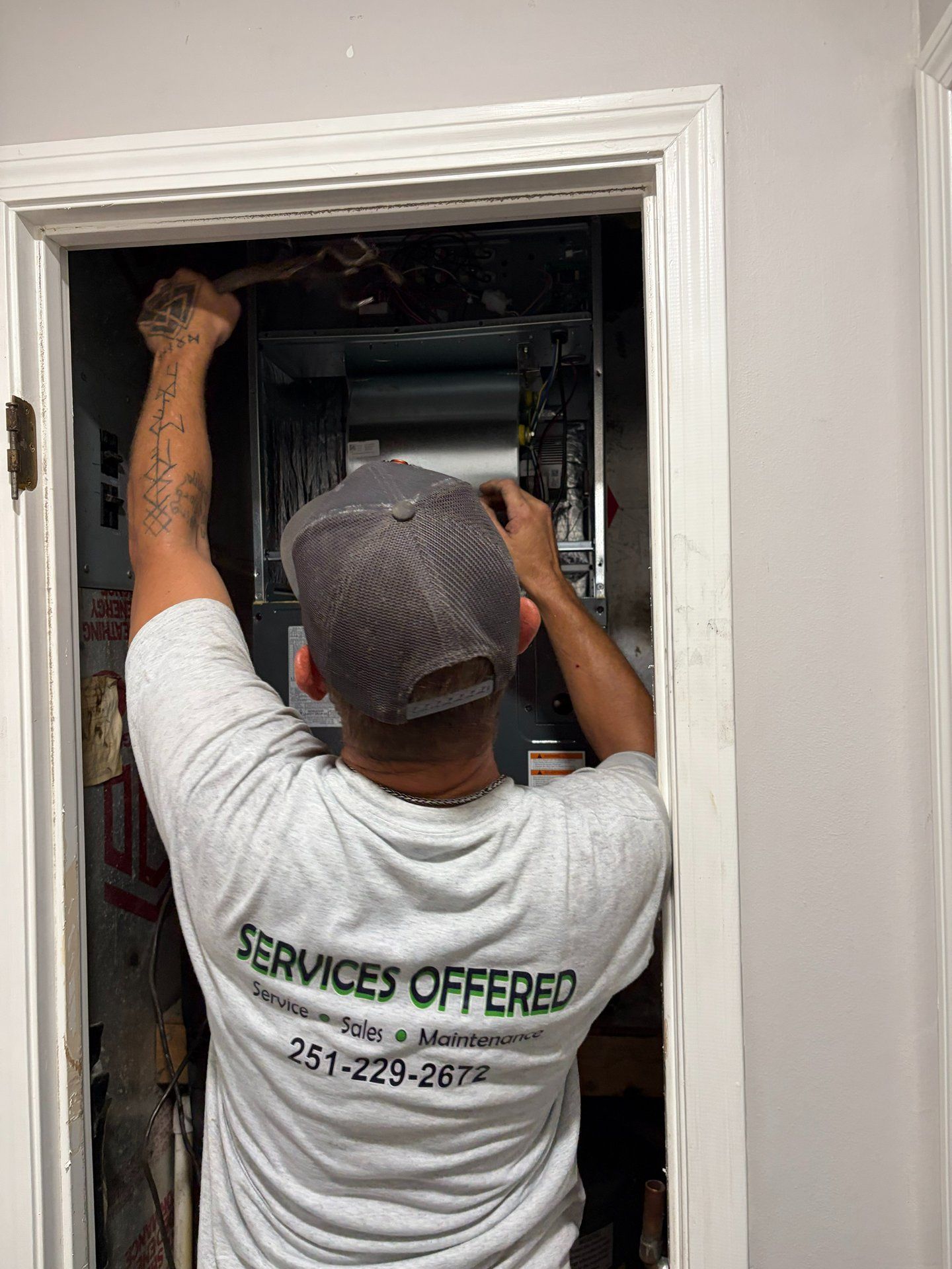 Worker reaching into an open wall panel inside a doorway, wearing a gray cap and white T-shirt.