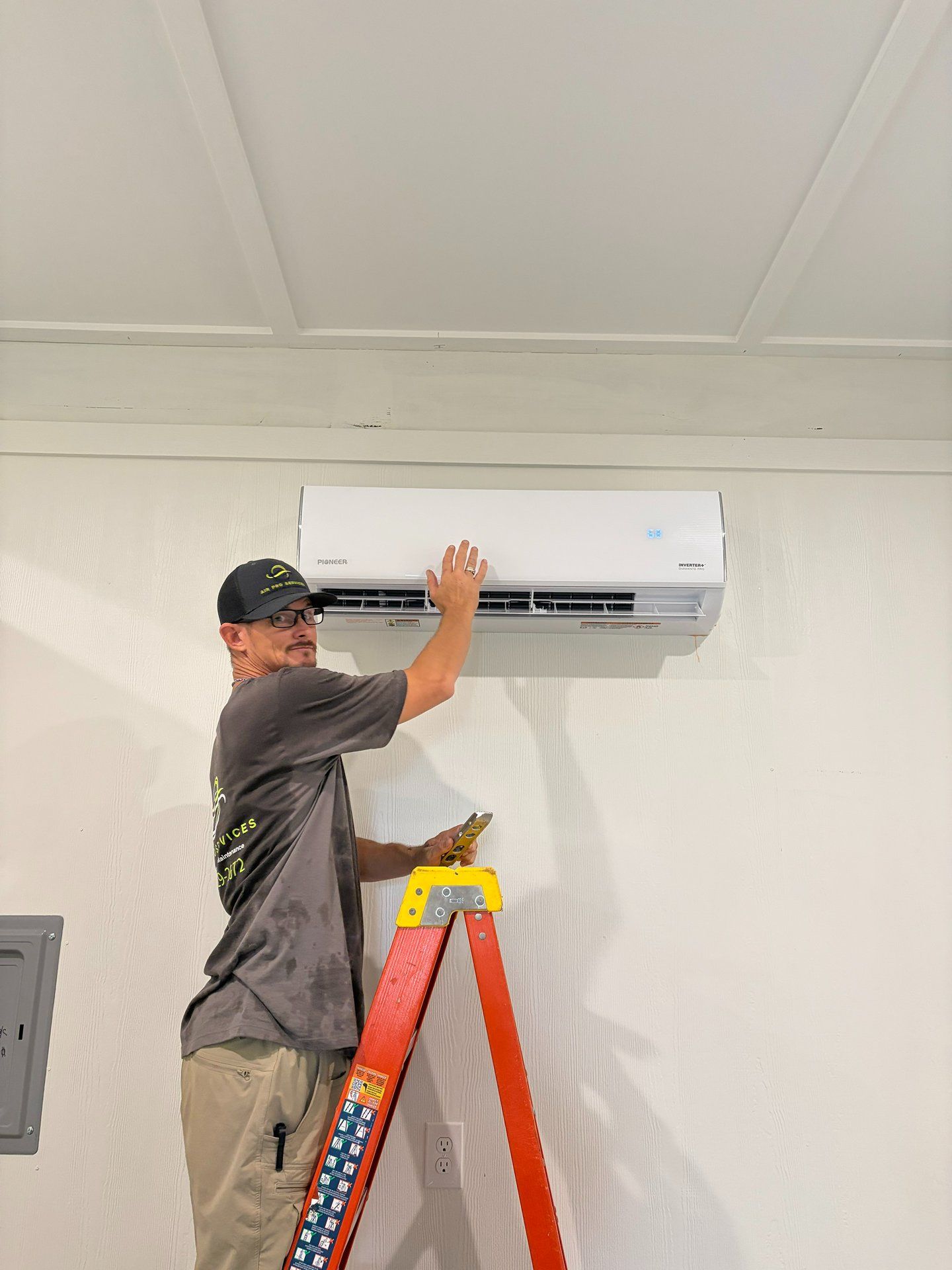 Worker installing a wall-mounted air conditioner on a ladder in a room with white walls.