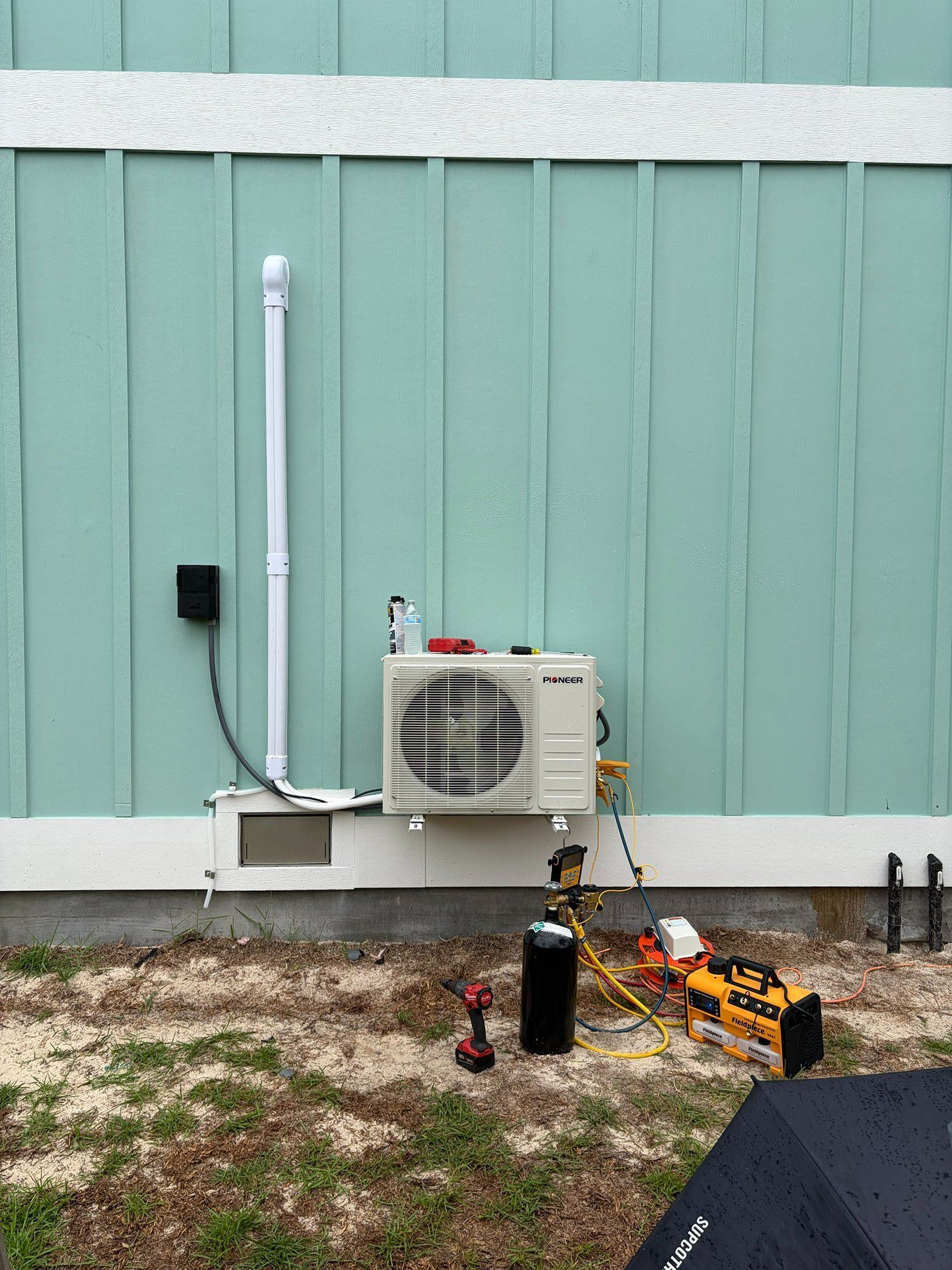 Outdoor HVAC unit mounted on a mint-green wall beside a black tank and tools on gravel.