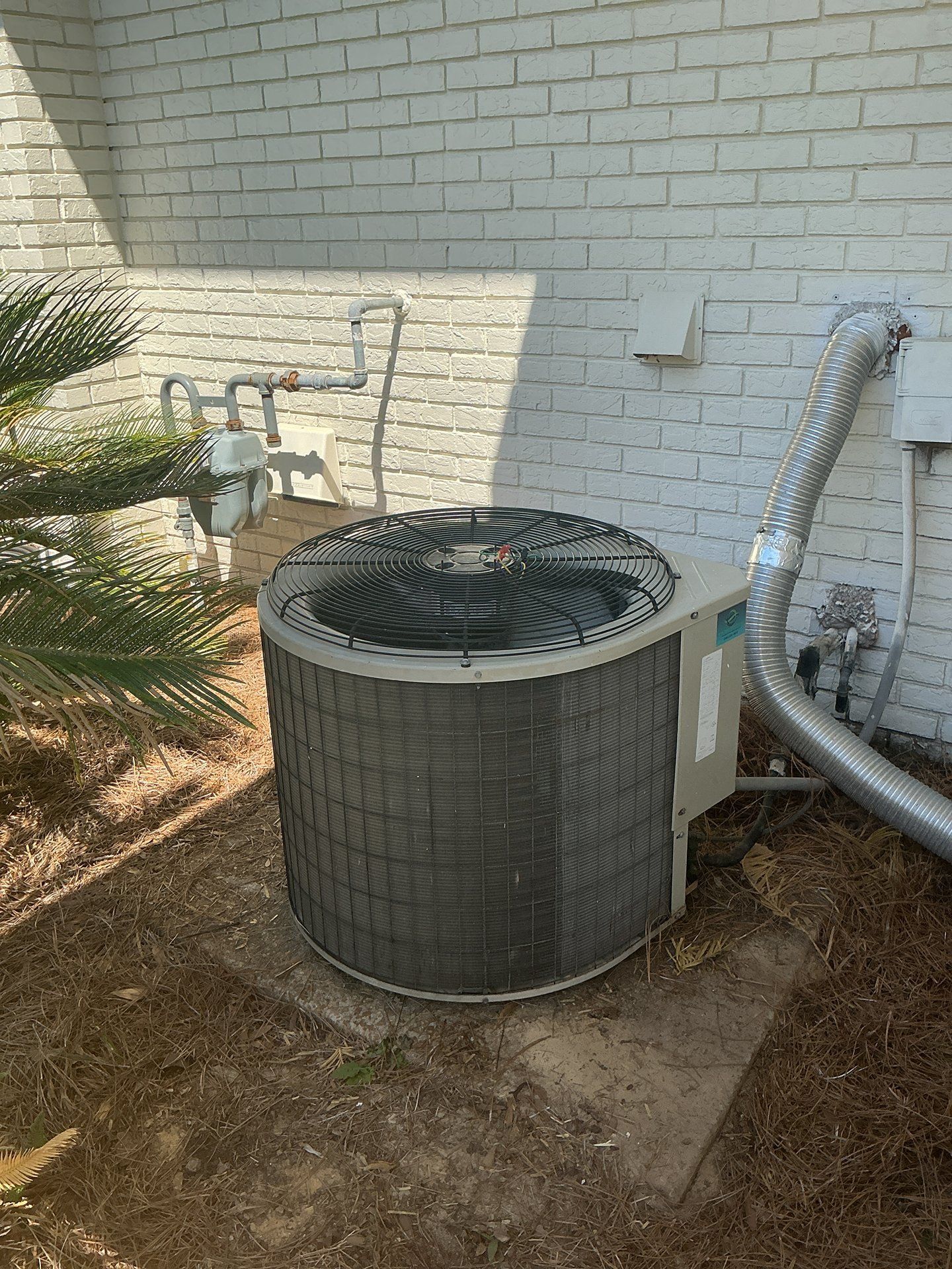 Outdoor air conditioner unit beside a brick house wall, with white pipes and a shaded fan top.