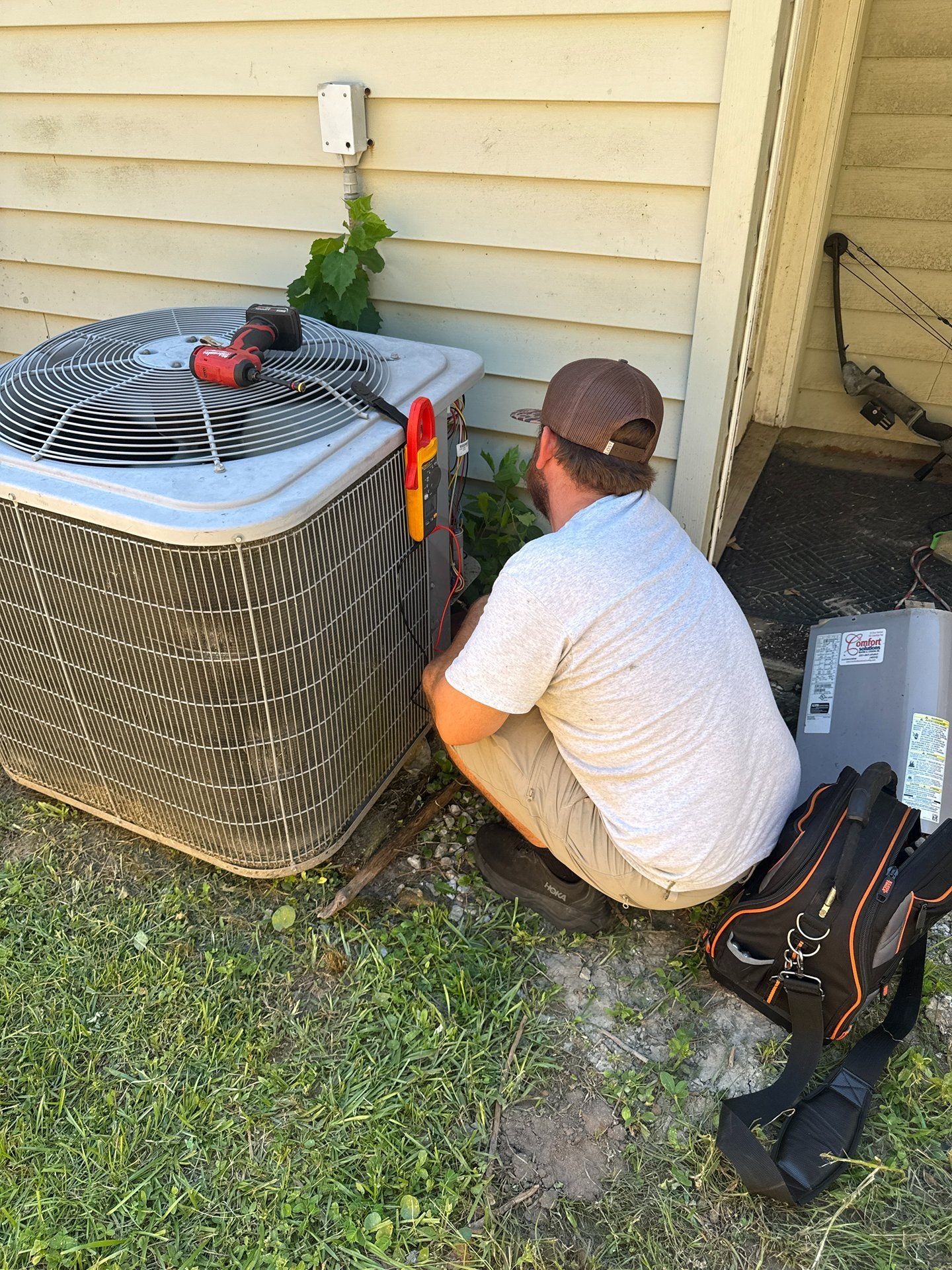 Technician kneeling beside outdoor air conditioner unit, inspecting equipment with tools nearby.