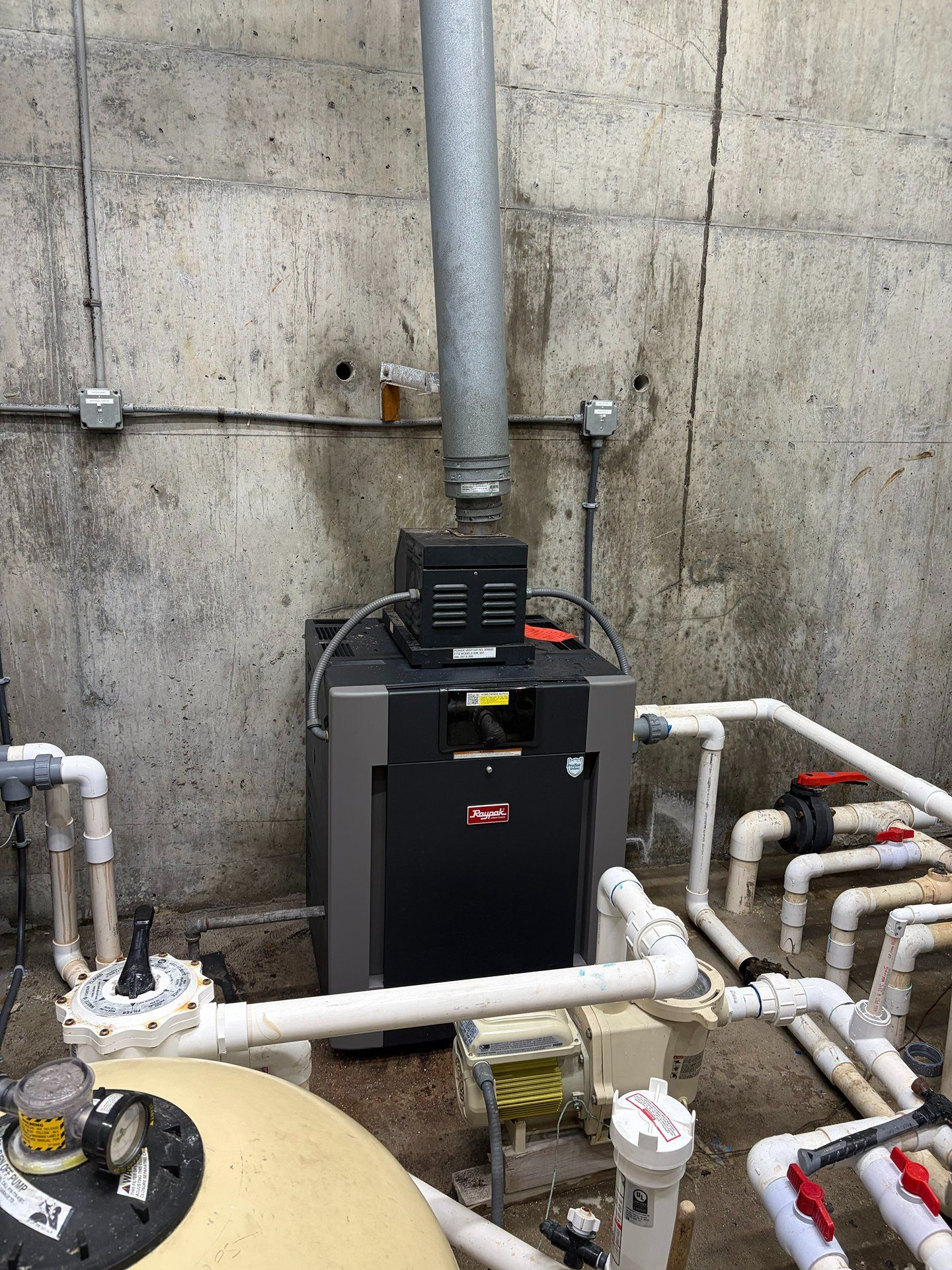 Industrial HVAC equipment and white pipes in a concrete basement room with a beige tank in the foreground