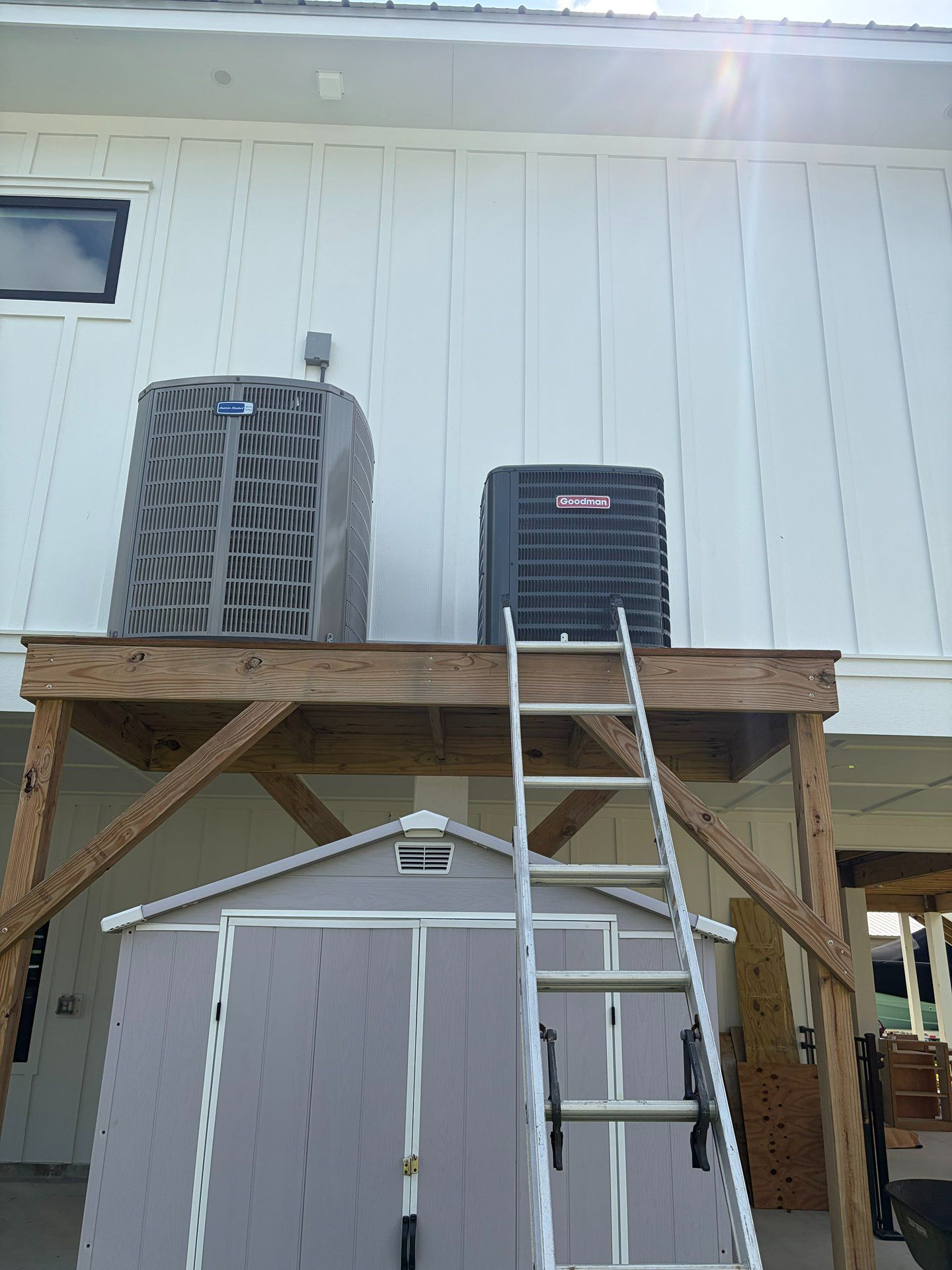 Two outdoor air conditioning units on a wooden platform with a ladder leaning below a white building