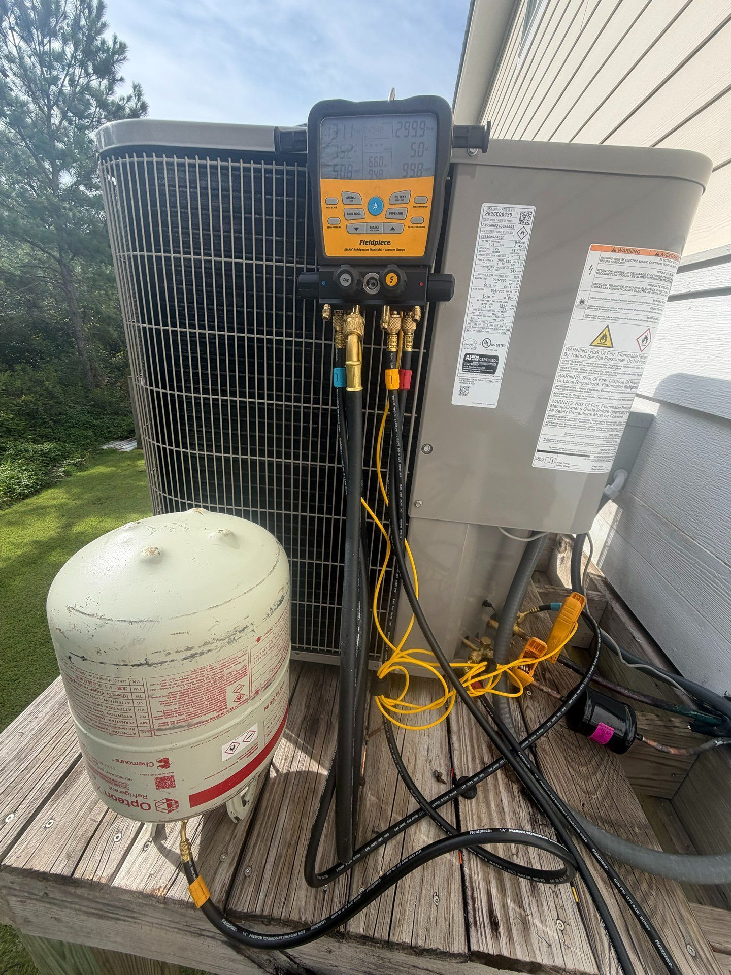 Outdoor HVAC unit with gauges, hoses, and a white refrigerant tank beside a wall.