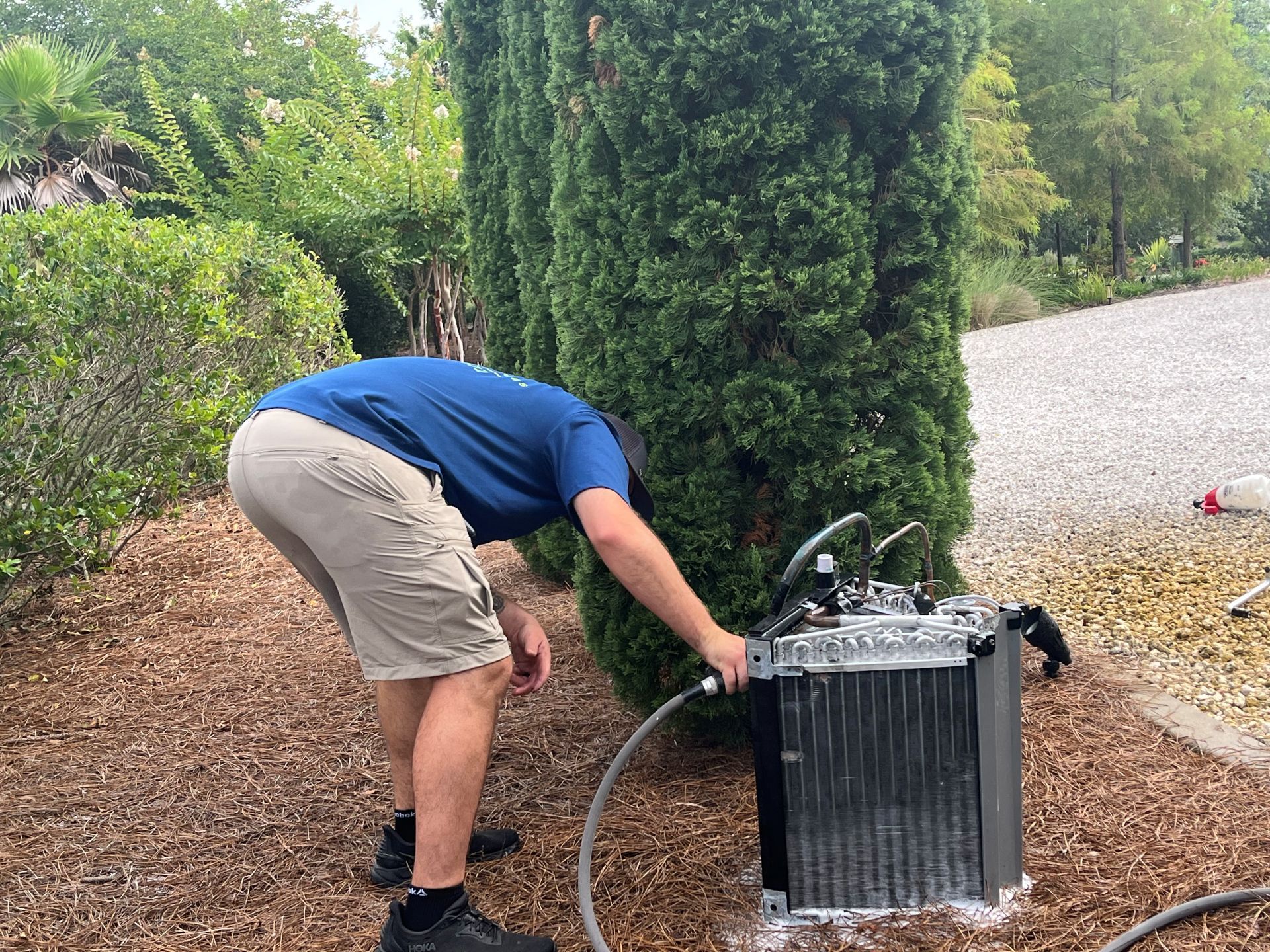 Person bending over an outdoor metal unit with a hose in a garden setting