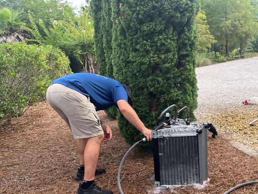 Person bending over a hose attached to a black metal box outdoors among gravel and trees