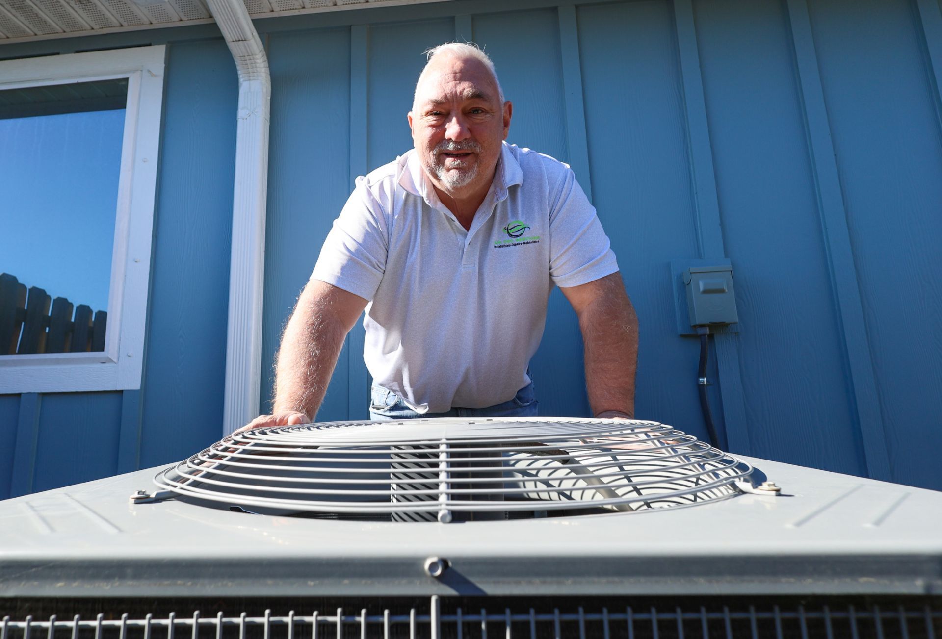 Man in white polo inspecting a rooftop HVAC unit beside a blue building.