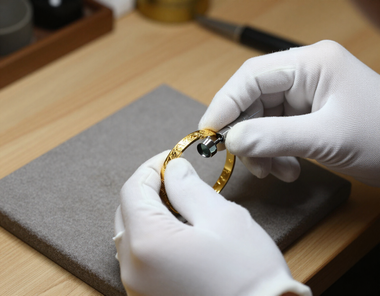 Behandschuhte Hände justieren eine goldene Armbanduhr auf einem grauen Kissen über einem Holztisch.