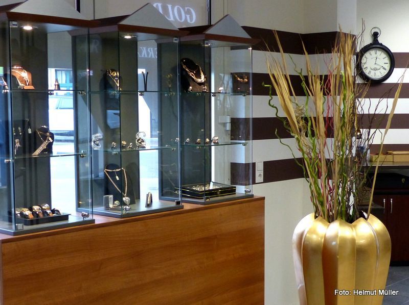 Vitrinen im Juweliergeschäft mit Uhren, einer großen goldenen Vase mit hohen Zweigen und einer Wanduhr auf der rechten Seite.
