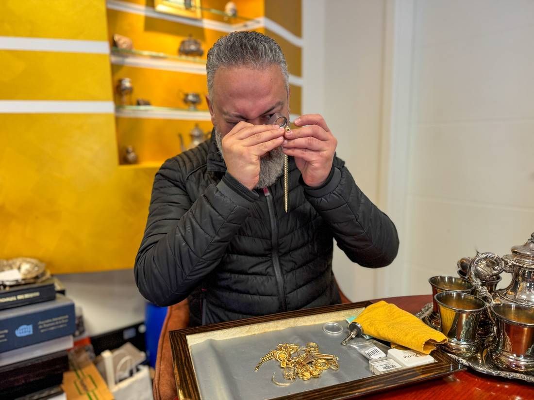 Eine Person sitzt in einem Innenraum und betrachtet einen kleinen Goldgegenstand auf einem Tisch mit Schmuckwerkzeugen und Tabletts.