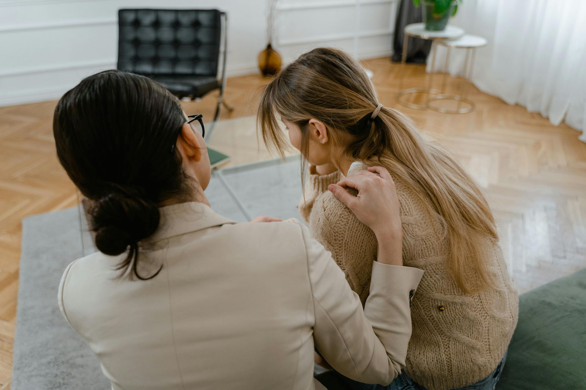 A person in a beige blazer comforts someone with long hair, placing a hand on their shoulder in a bright, modern room.