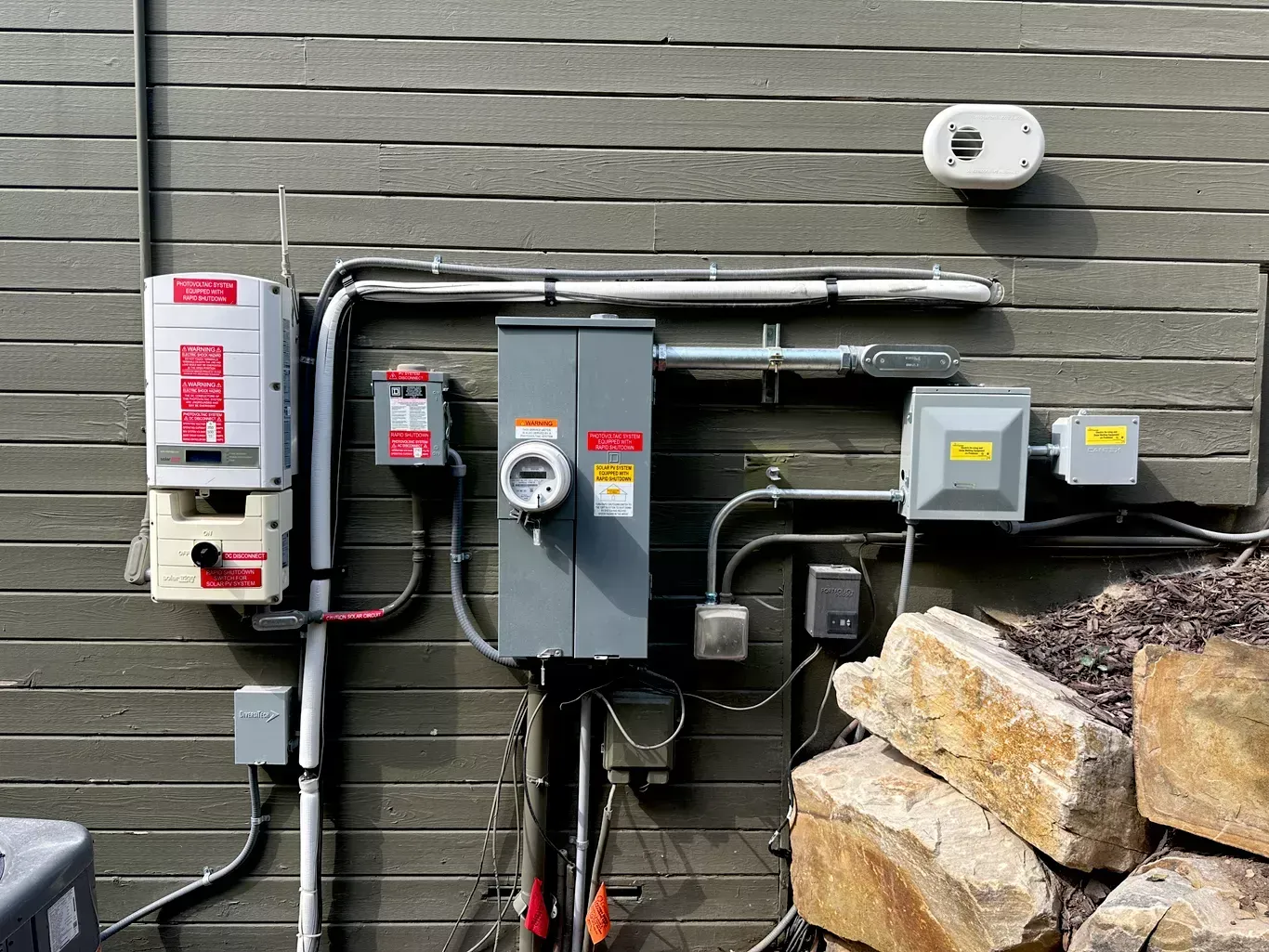 Electrical components, including a meter, breaker boxes, and conduit, mounted on a gray wooden wall.