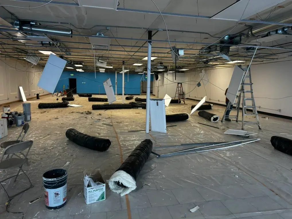 Construction site: large room with debris, rolls of black tubing, and white panels on the floor and walls.