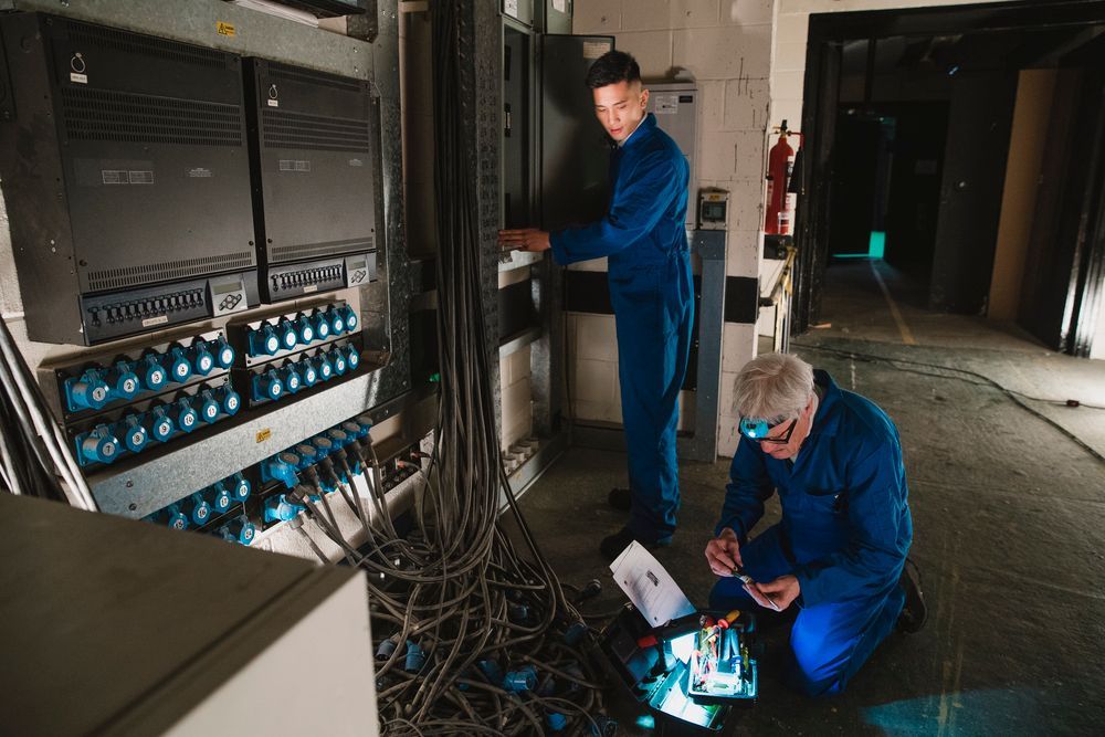 Two technicians in blue coveralls inspect electrical equipment in a dim commercial space.