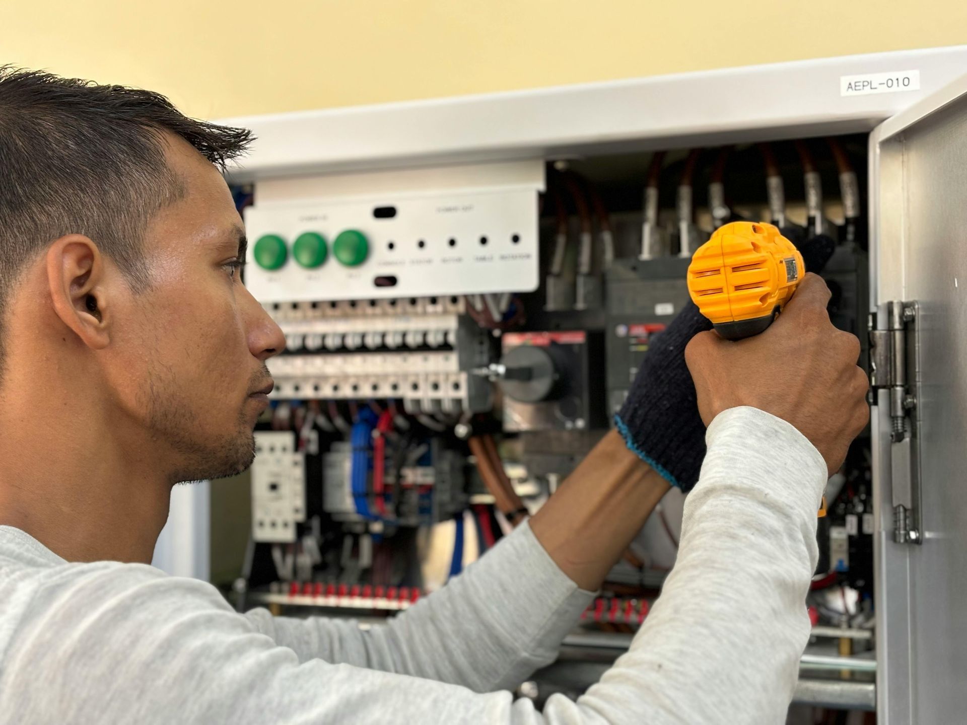 Man working on electrical panel, holding a yellow tool, wearing grey gloves, inside a room.