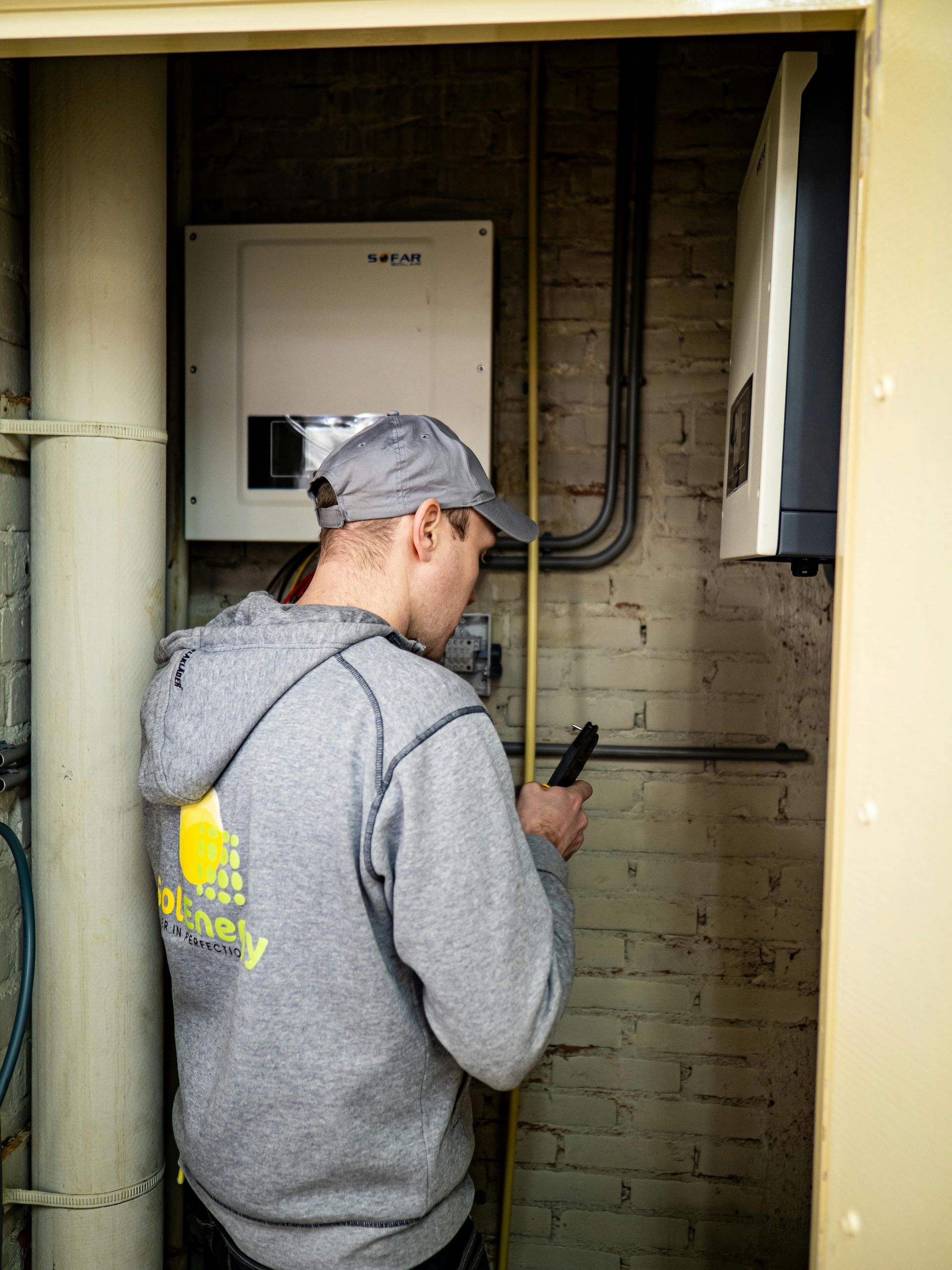 Man in gray hoodie examines electrical equipment in a brick-lined room.