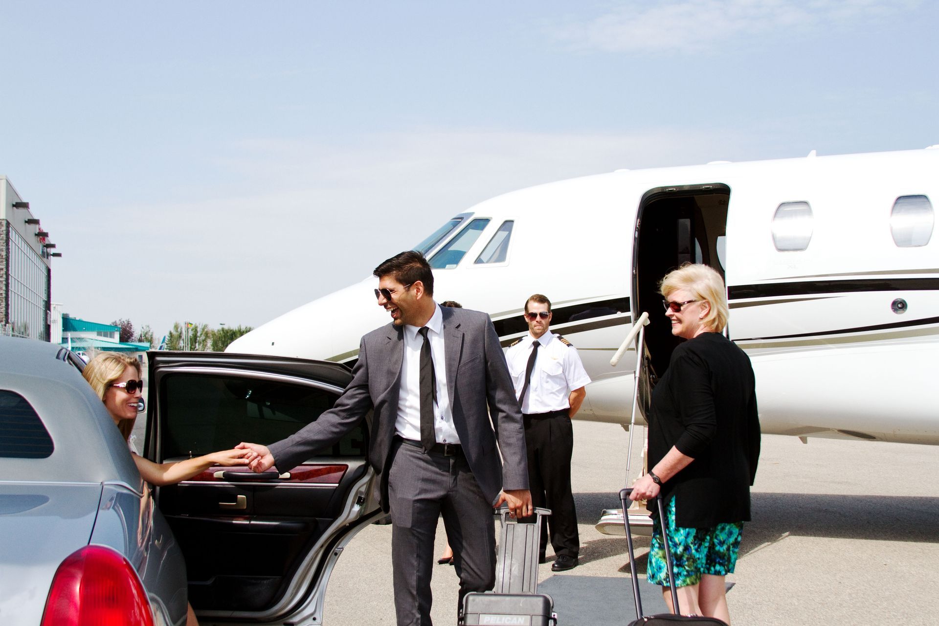 A man in a suit assists a woman exiting a car near a private jet; another woman waits with luggage. A man in a suit assists a woman exiting a car near a private jet; another woman waits with luggage.