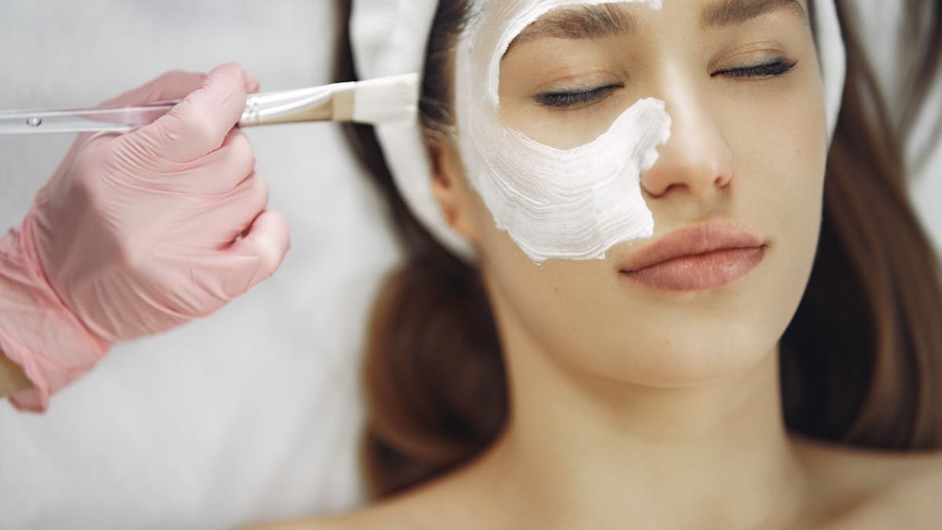 Aplicación de una mascarilla facial en el rostro de una mujer con una brocha y una mano enguantada en un entorno de spa.