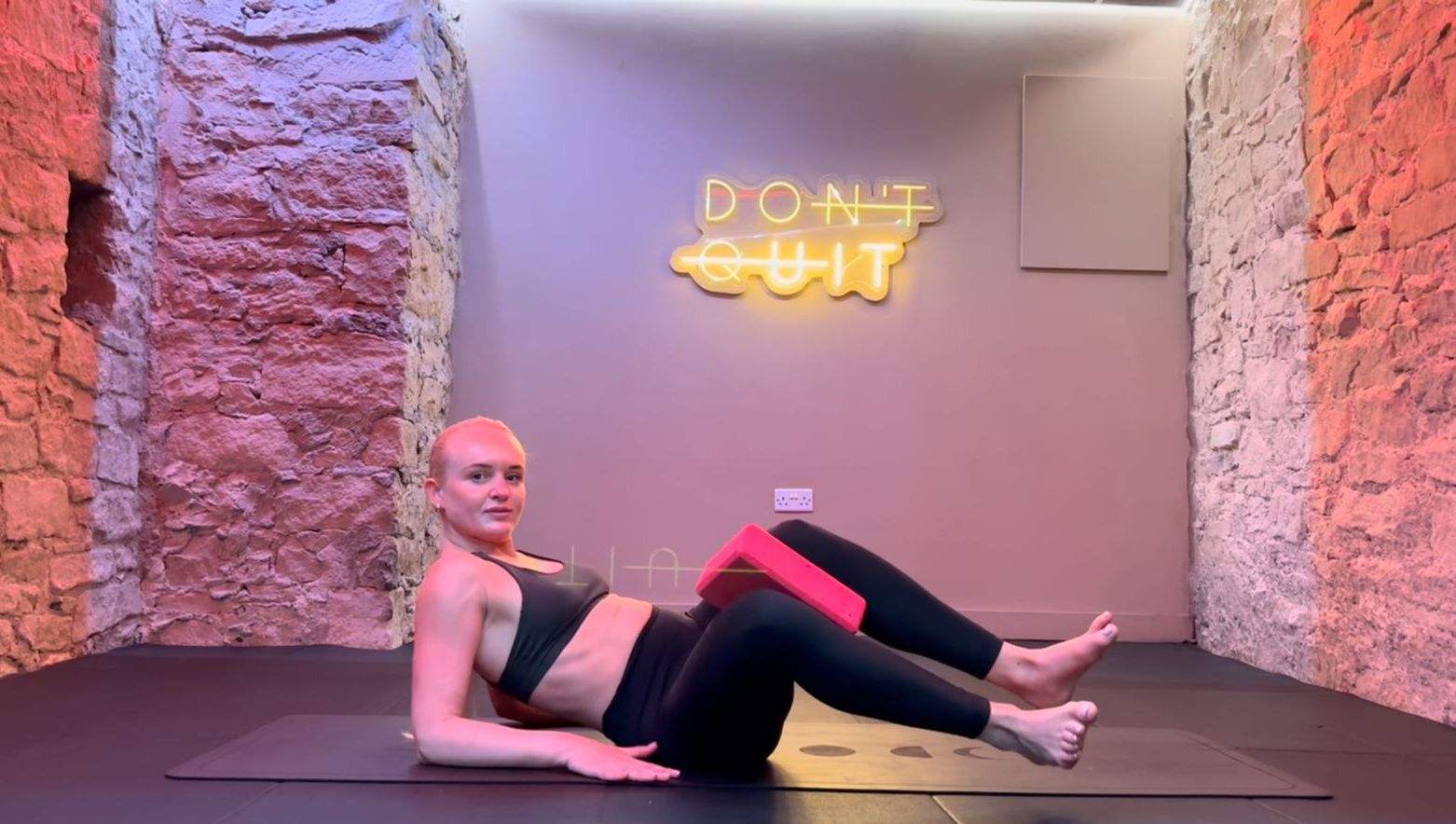 Woman in fitness attire performs glute bridge exercise on a mat, holding a block between her knees.