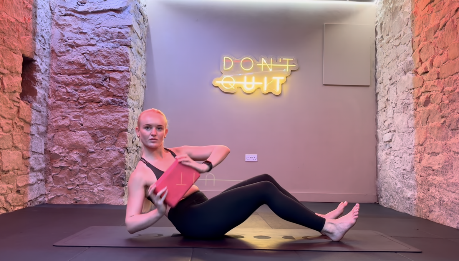 Woman exercising, seated on mat, holding a block and twisting torso. The words 