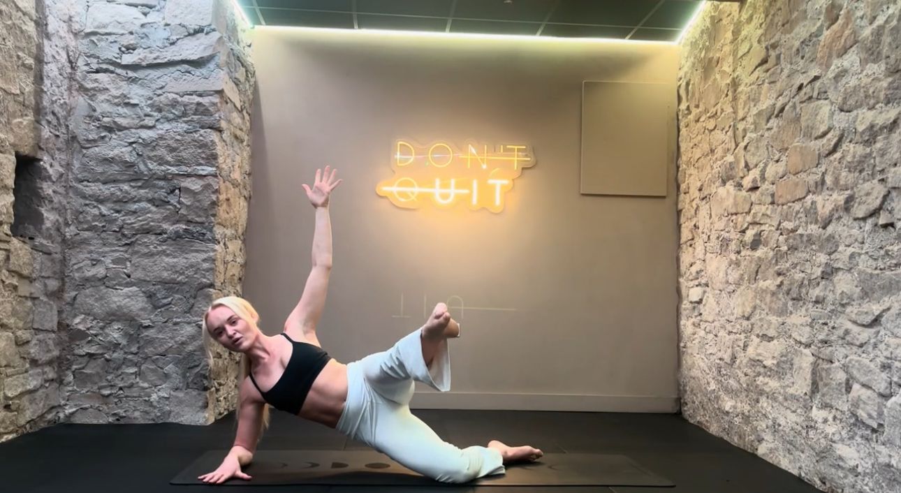 Woman in yoga pose on a mat with arm raised, leg bent. Brick wall backdrop, neon sign says