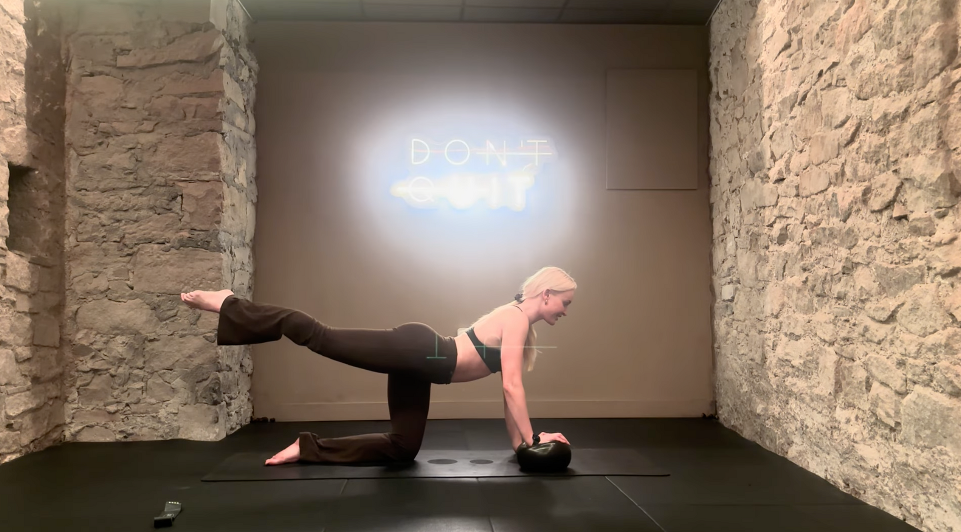 Woman doing an ab exercise on a mat, raising one leg and arm, in a room with stone walls and a 