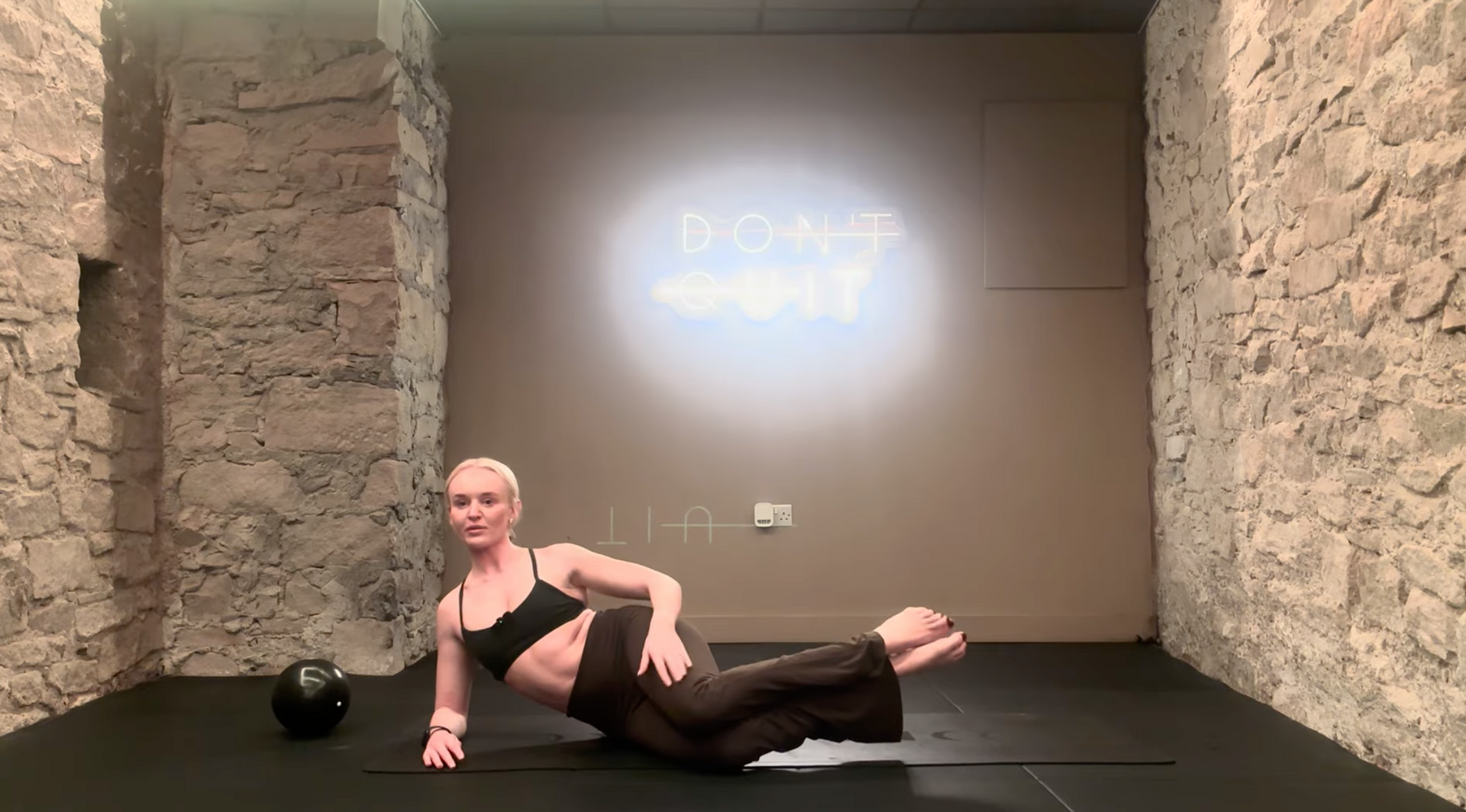 Woman doing an ab exercise on a mat, raising one leg and arm, in a room with stone walls and a 
