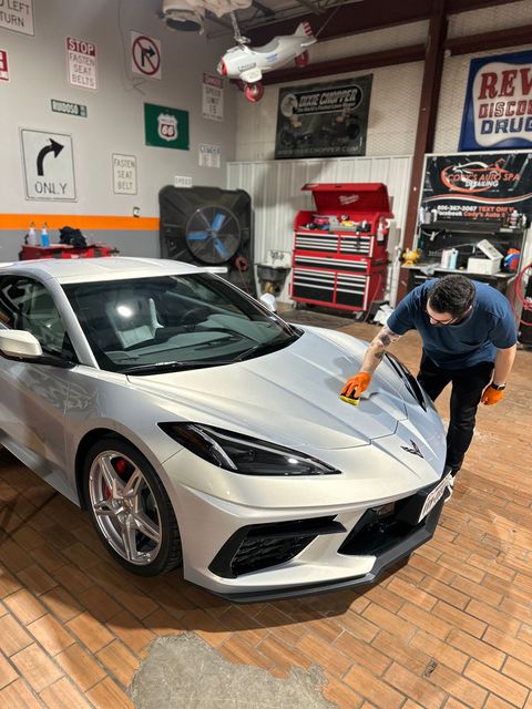 A person wearing orange gloves polishes the silver hood of a sports car in a decorated garage.