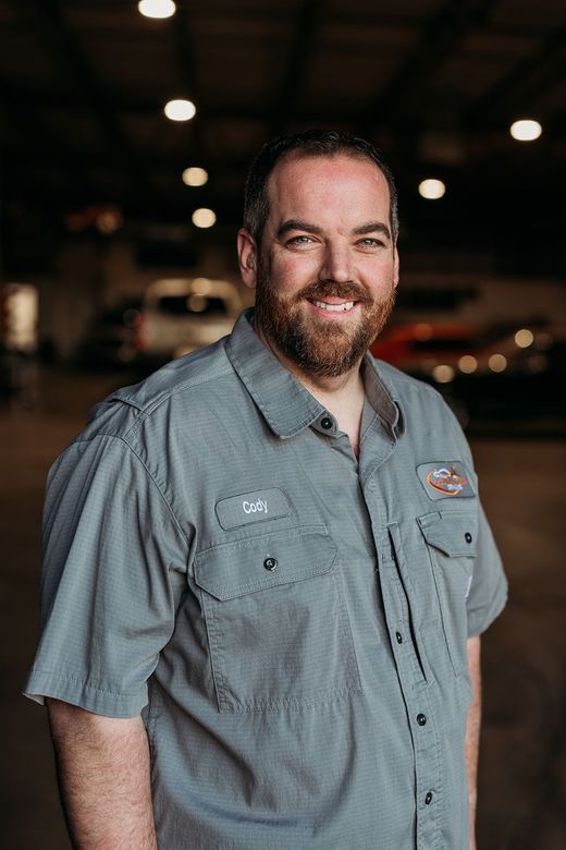 A person with a beard and glasses in a gray T-shirt with tattoos on their arm, standing in a dimly lit garage.