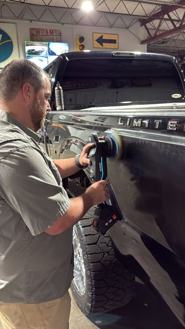 A person uses an electric buffer to polish the black side panel of a large pickup truck in a workshop.