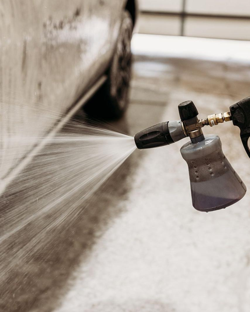 A foam cannon sprays a stream of soap onto the side of a dirty car.