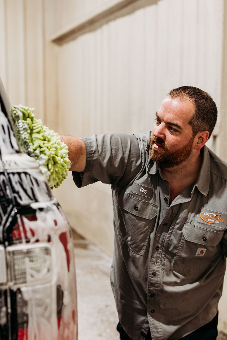 A person in a gray shirt uses a green mitt to wash a soapy vehicle in a garage.