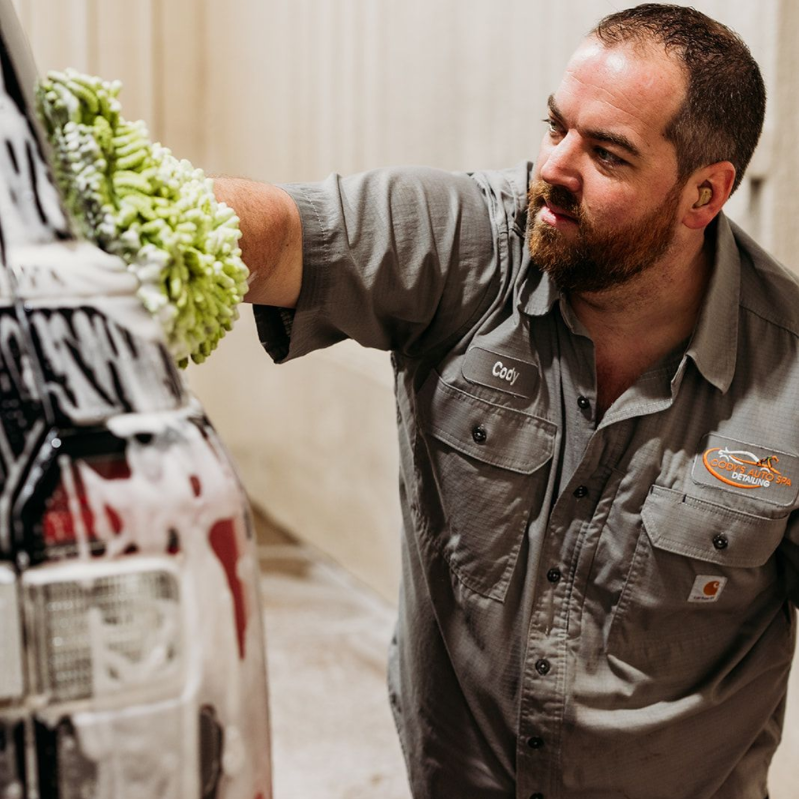 A person in a gray work shirt uses a green wash mitt to clean a car covered in soap suds in an indoor bay.