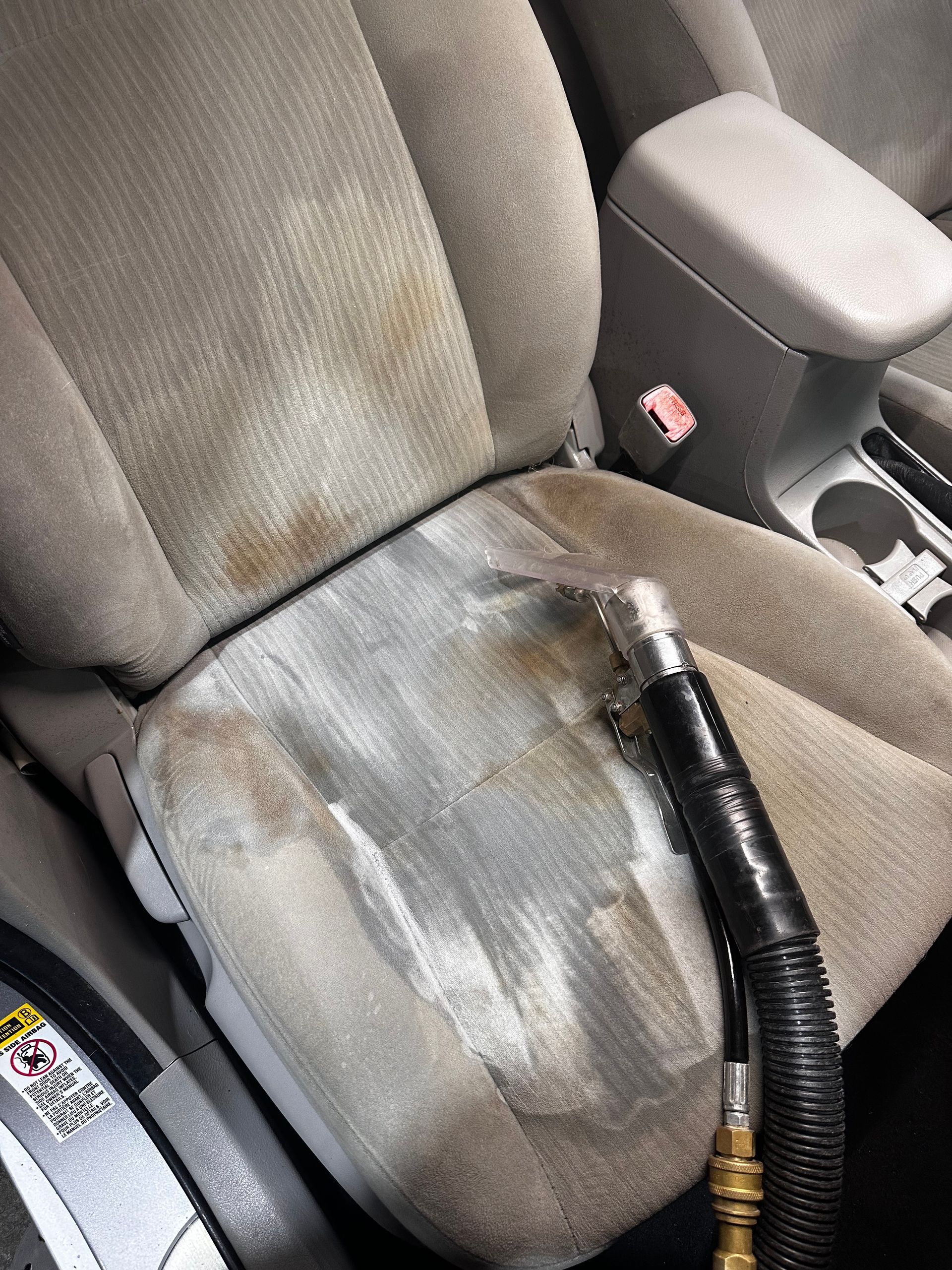 A car upholstery extractor tool cleaning a stained, light-colored fabric seat, showing a contrast between dirty and clean.
