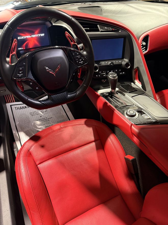 Interior of a Chevrolet Corvette featuring a black steering wheel, red perforated leather seats, and a dashboard screen.