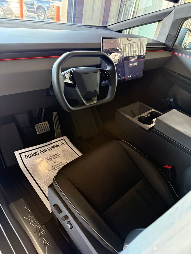 Interior of a Tesla Cybertruck with a futuristic steering yoke, large center touchscreen, and black seat.
