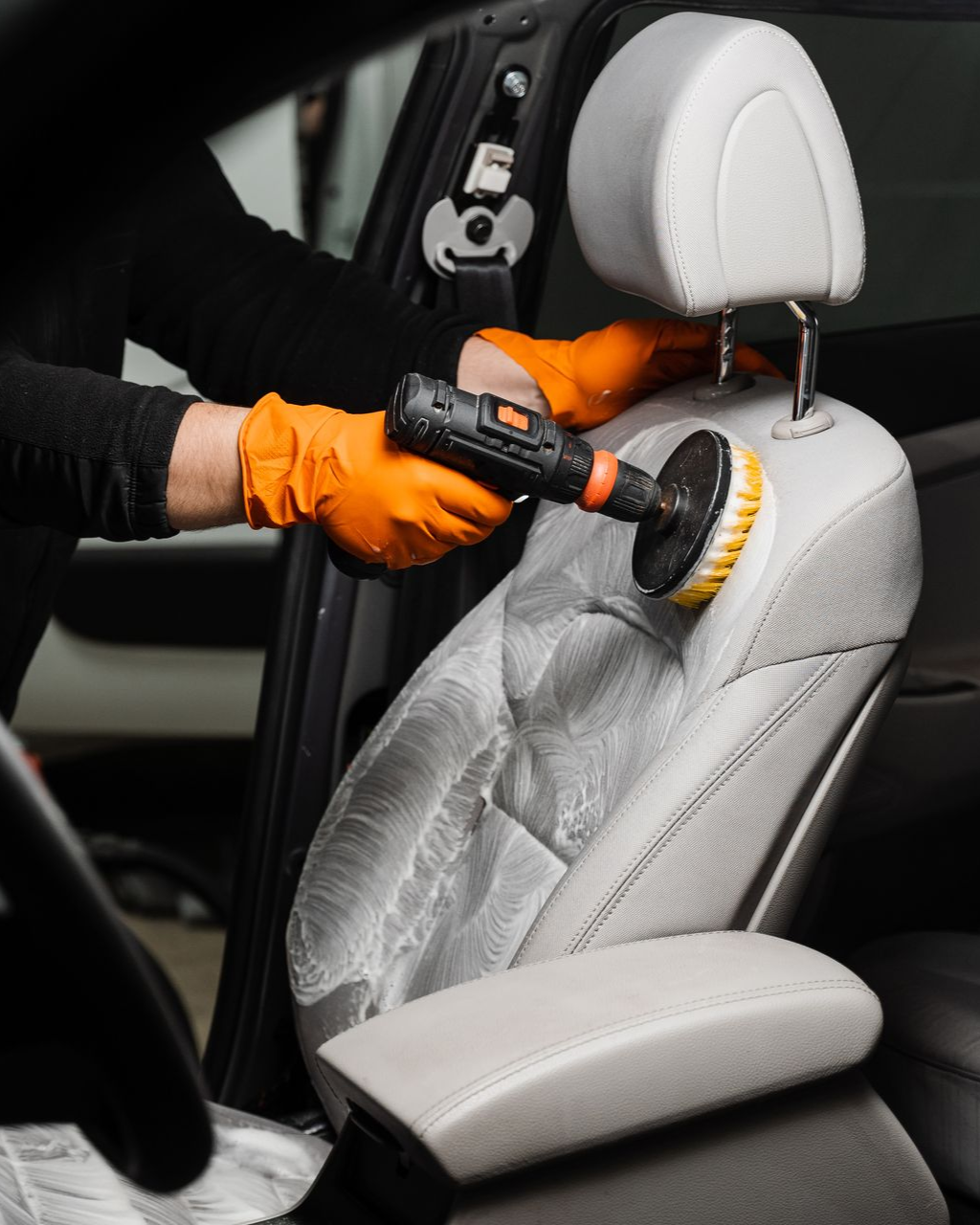 A person wearing orange gloves uses a power drill with a circular brush attachment to deep clean a light grey car seat.