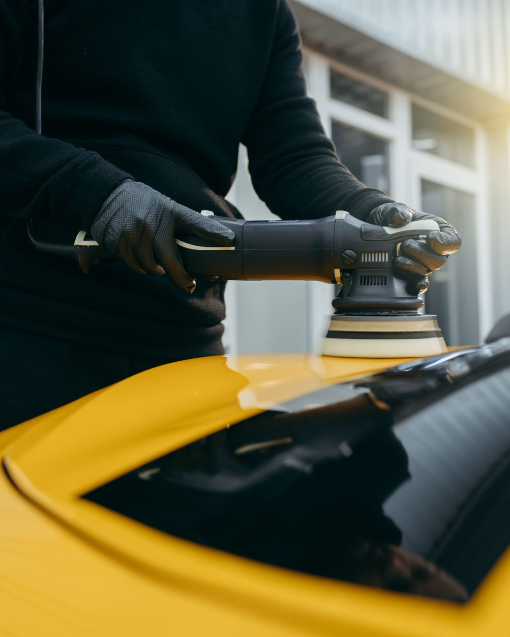 A person in black gloves uses an electric buffer to polish the yellow hood of a car.