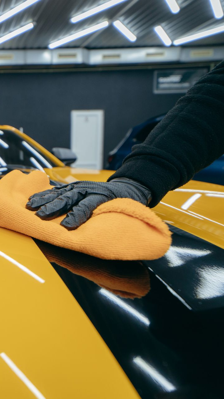 A person wearing a black glove wipes the glossy yellow hood of a car with an orange microfiber cloth in a studio.