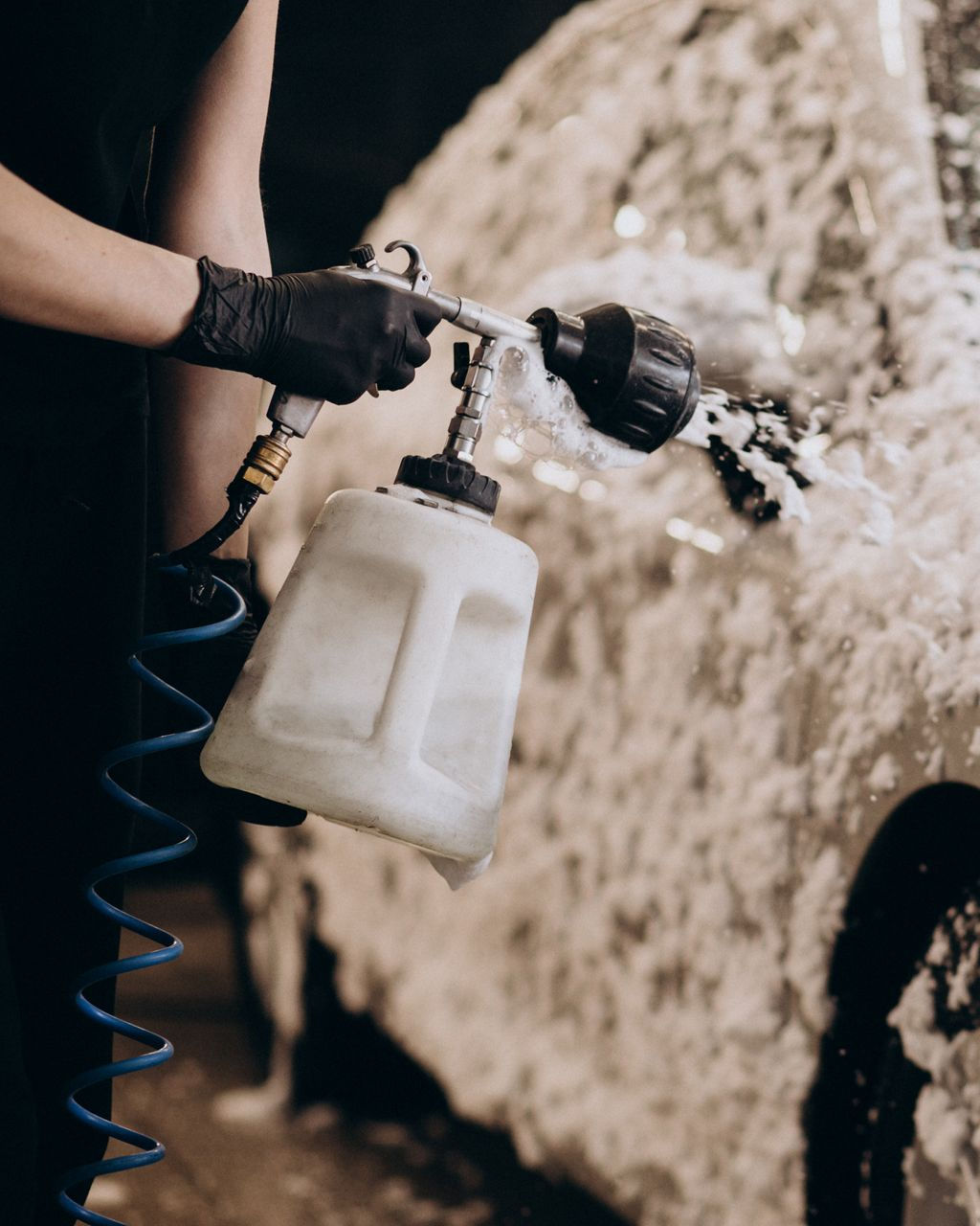 A person wearing black gloves uses a pneumatic spray gun to coat a vehicle with thick, white cleaning foam.