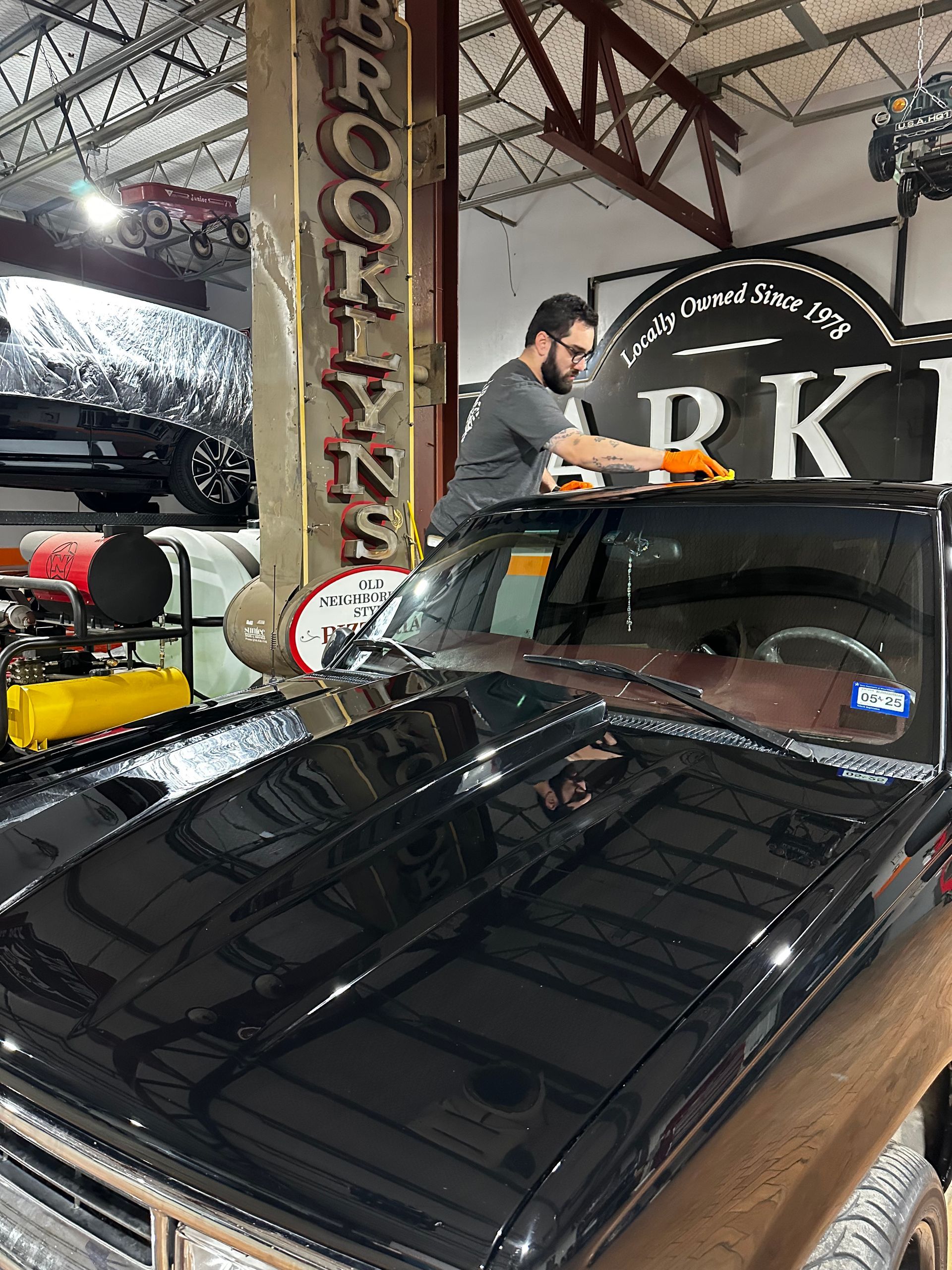 A person wearing orange gloves cleans the windshield of a shiny black vehicle inside an automotive shop.