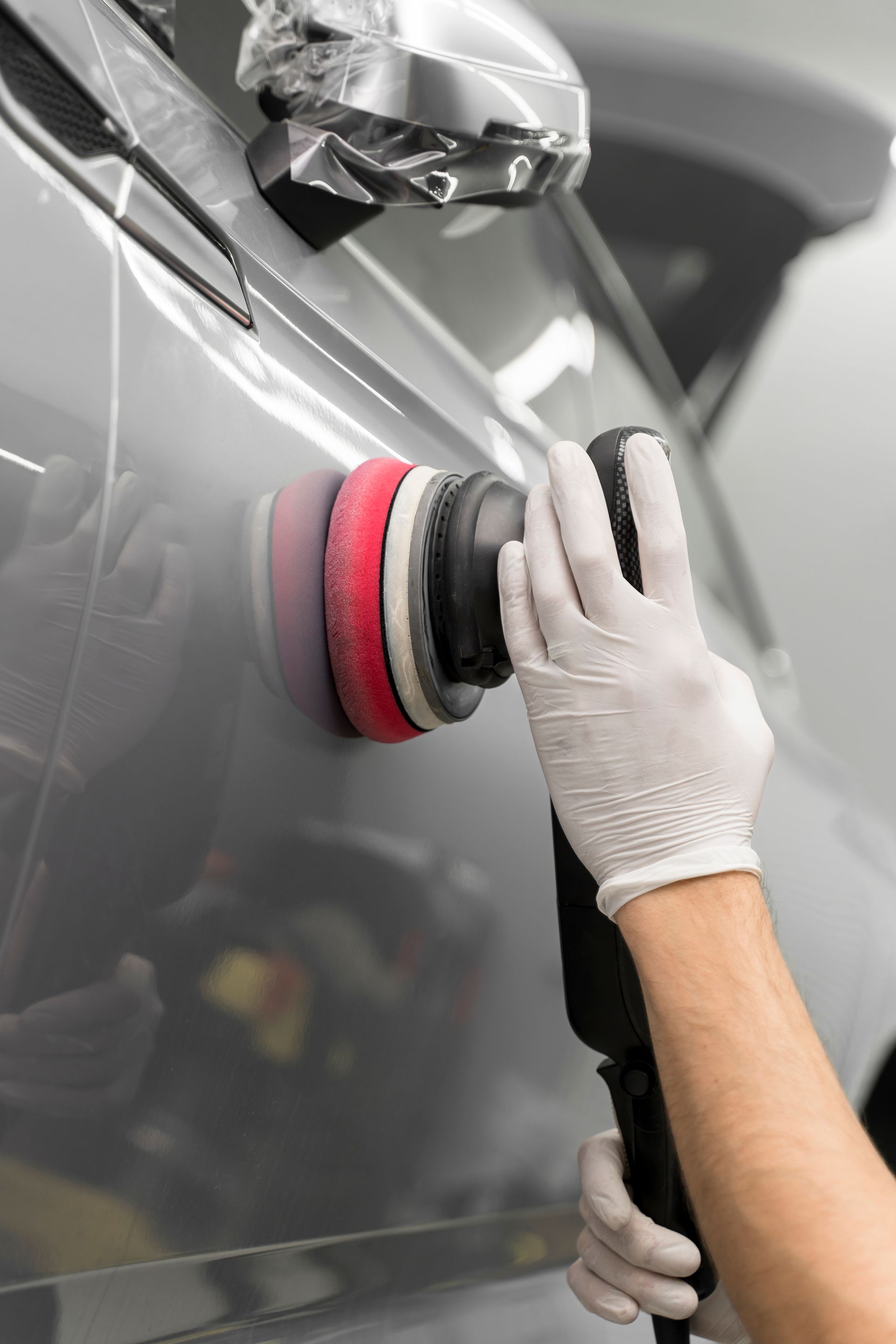 A gloved hand uses a rotary polisher with a red buffing pad on a grey car door.