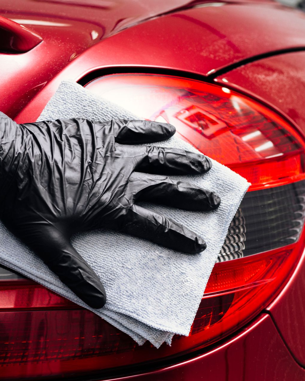 A hand in a black nitrile glove wipes a red car's taillight with a grey microfiber cloth.